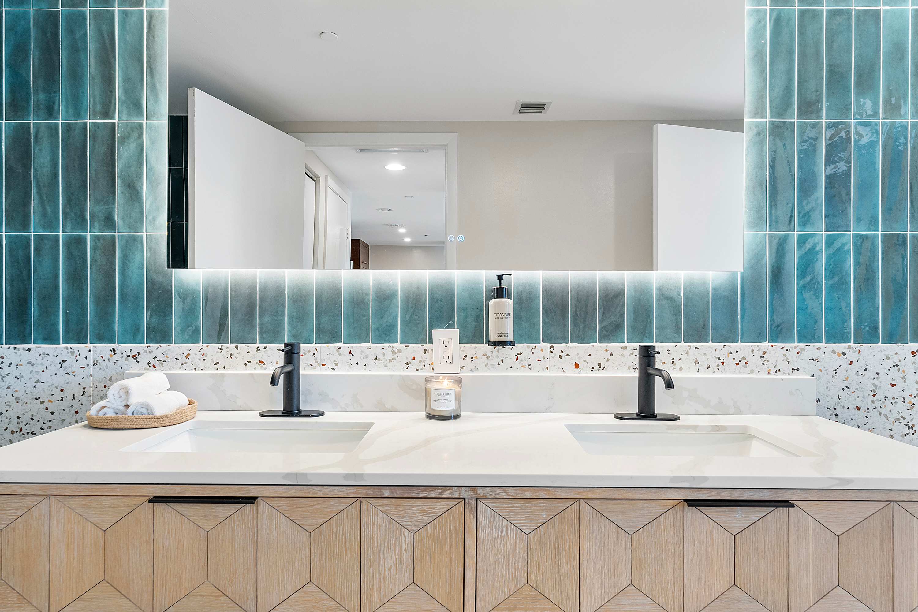 Close-up of a stylish double vanity in a modern bathroom, showcasing light wood cabinetry with geometric patterns, white marble countertops, and black faucets. An illuminated mirror is above teal subway tiles.