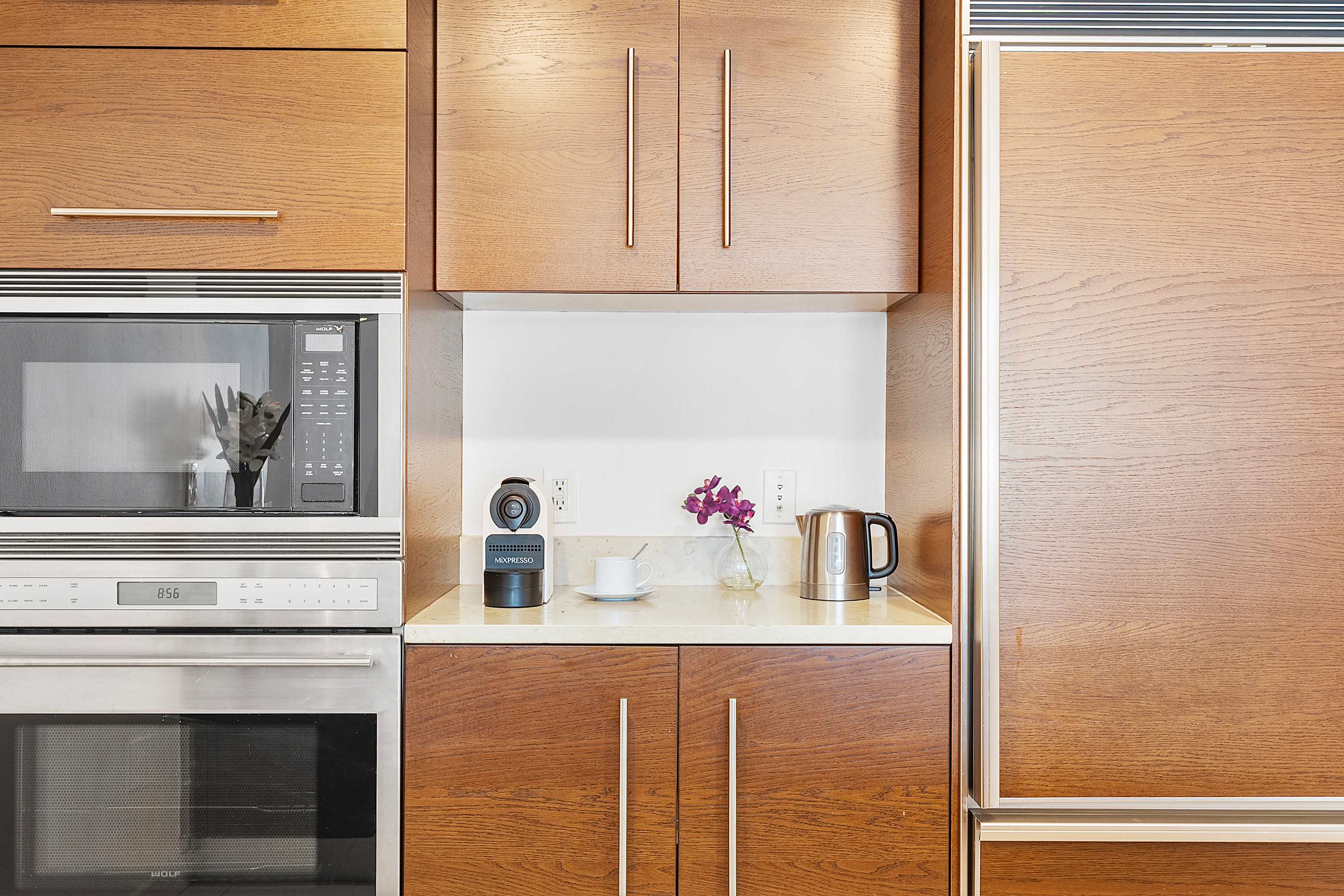 Close-up of a modern kitchen counter with built-in stainless steel microwave and oven, a coffee machine, and an electric kettle. Light wood cabinetry and a white backsplash complete the sleek design.