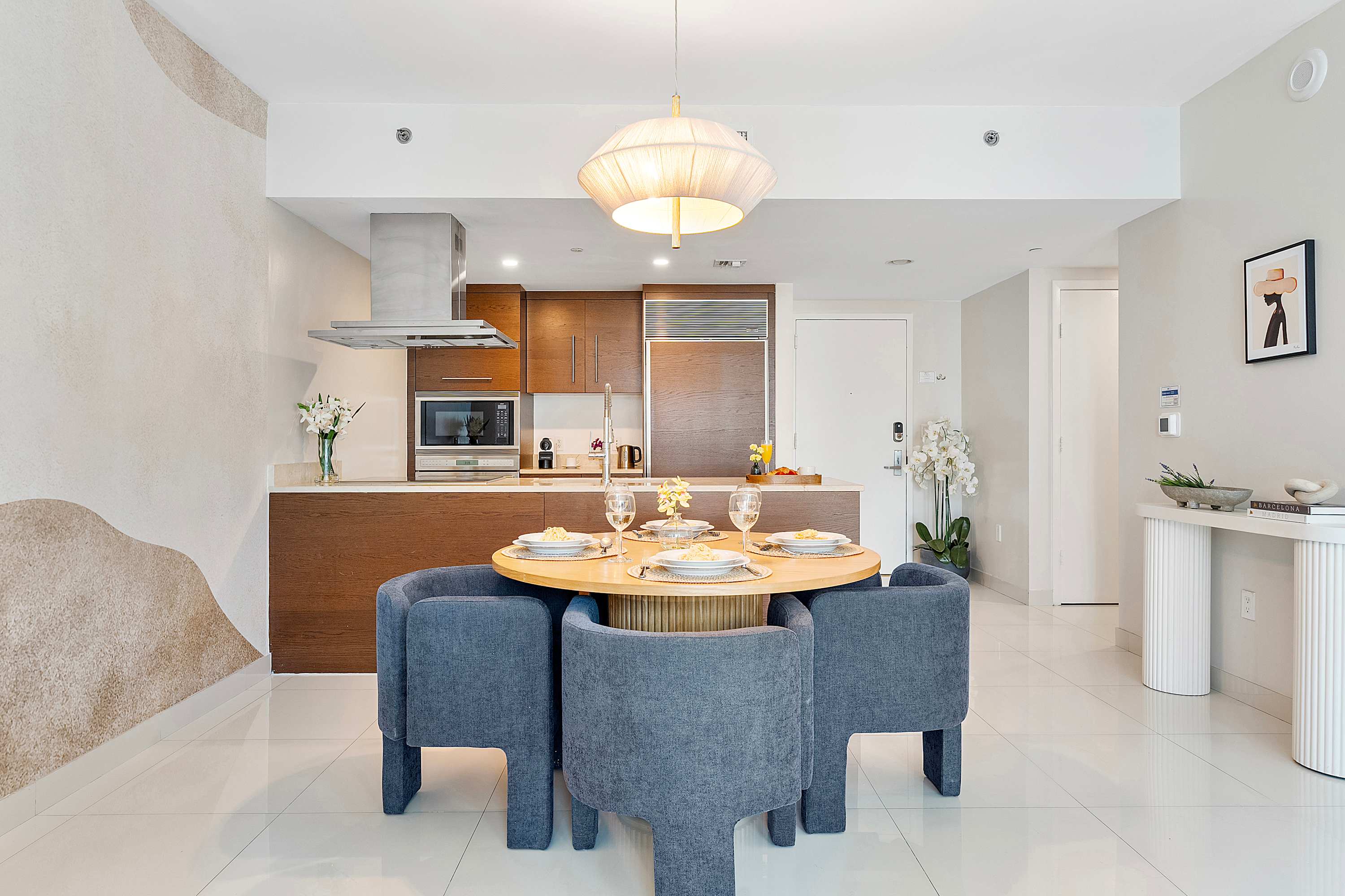 Contemporary kitchen and dining area featuring dark wood cabinetry, stainless steel appliances, a light wood round dining table with blue upholstered chairs, and a decorative accent wall.