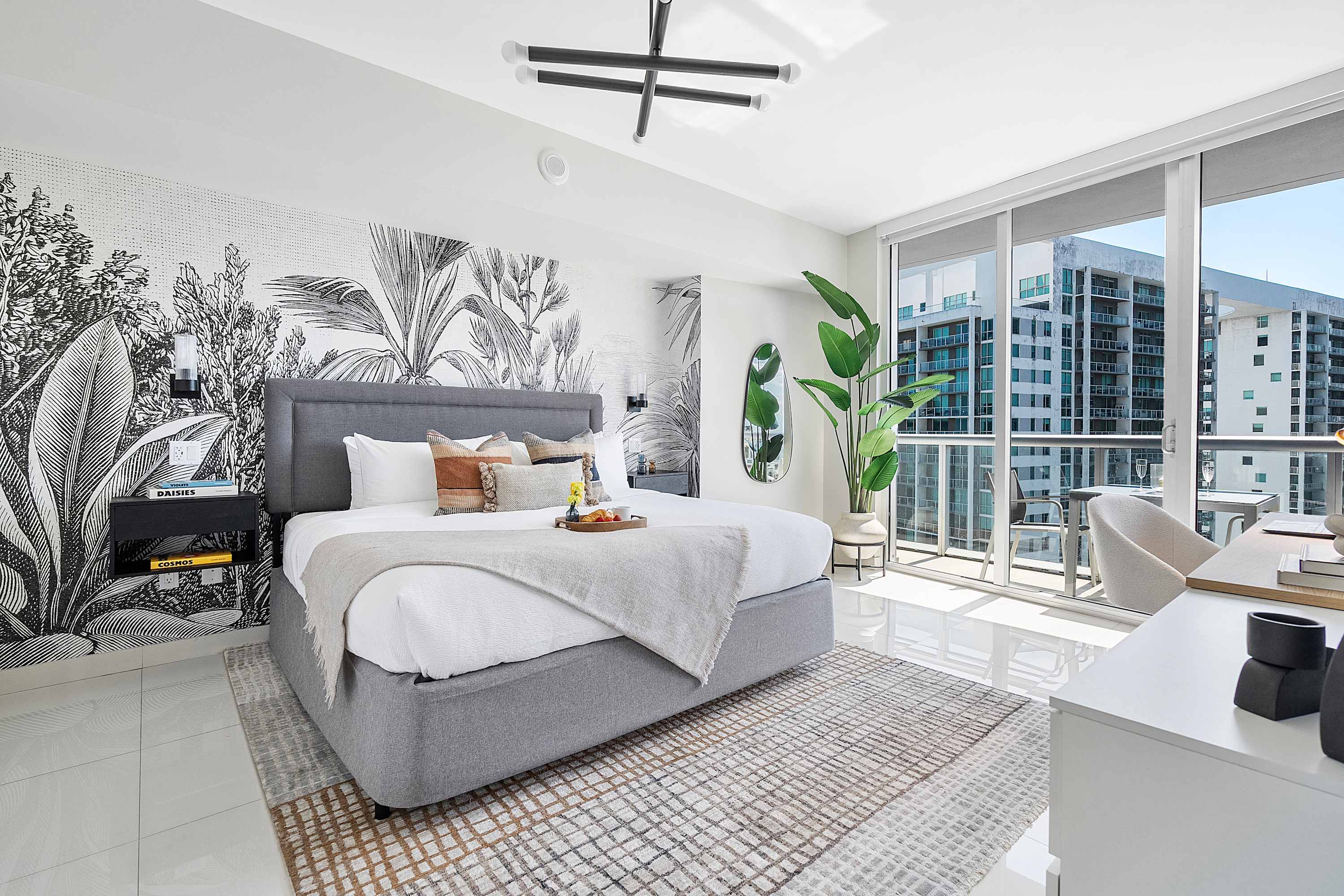 Bright bedroom with a gray upholstered king-size bed, a patterned rug, and a large window offering city views. A black and white botanical mural covers the wall behind the bed, complemented by a modern chandelier.