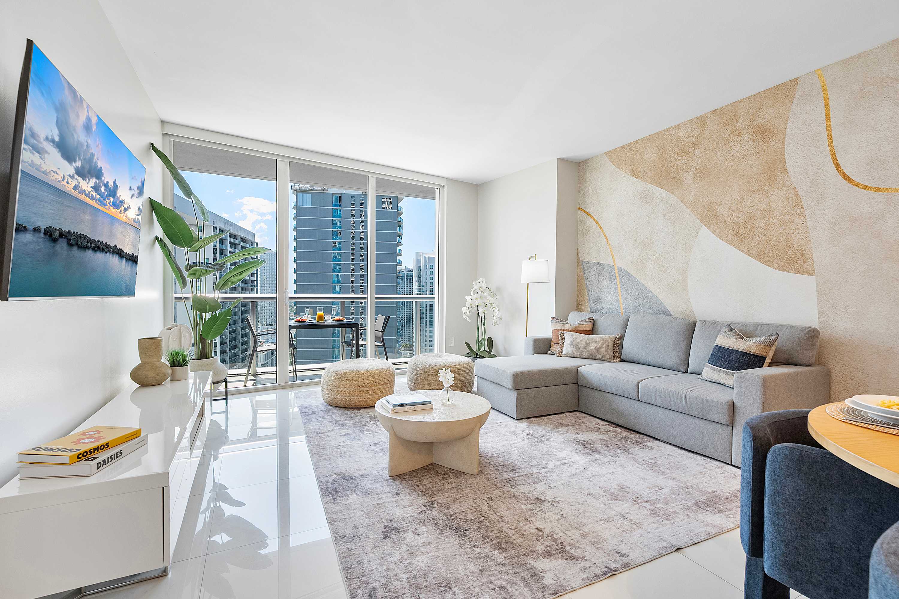 Bright living room with a grey sectional sofa, a round coffee table, and a large flat-screen TV, featuring a decorative accent wall and floor-to-ceiling windows offering city views and balcony access.