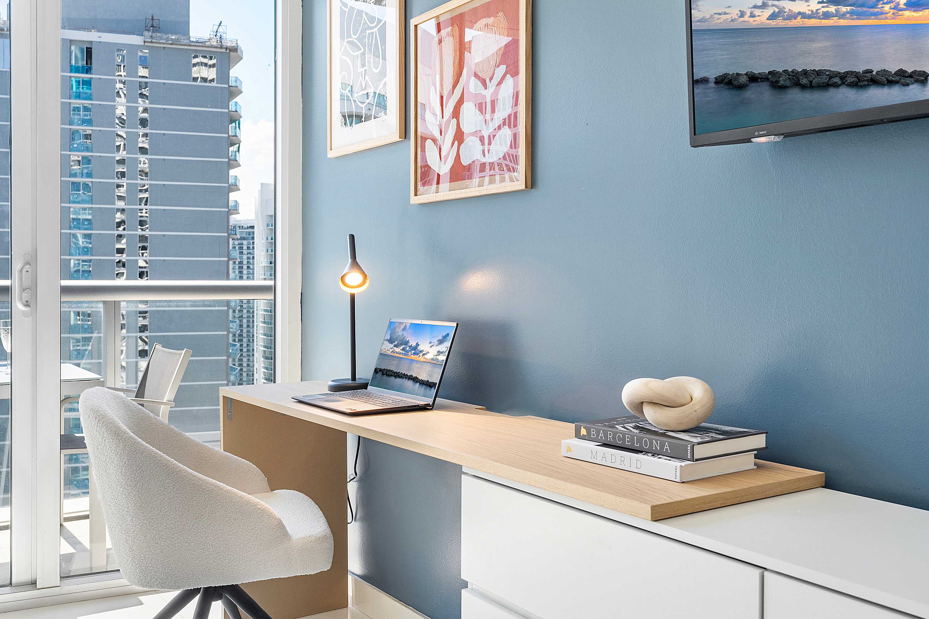 Modern workspace with a light wood desk, a white swivel chair, and a laptop. The area features a blue accent wall, framed art, and a large window with city views, creating a bright and functional nook.