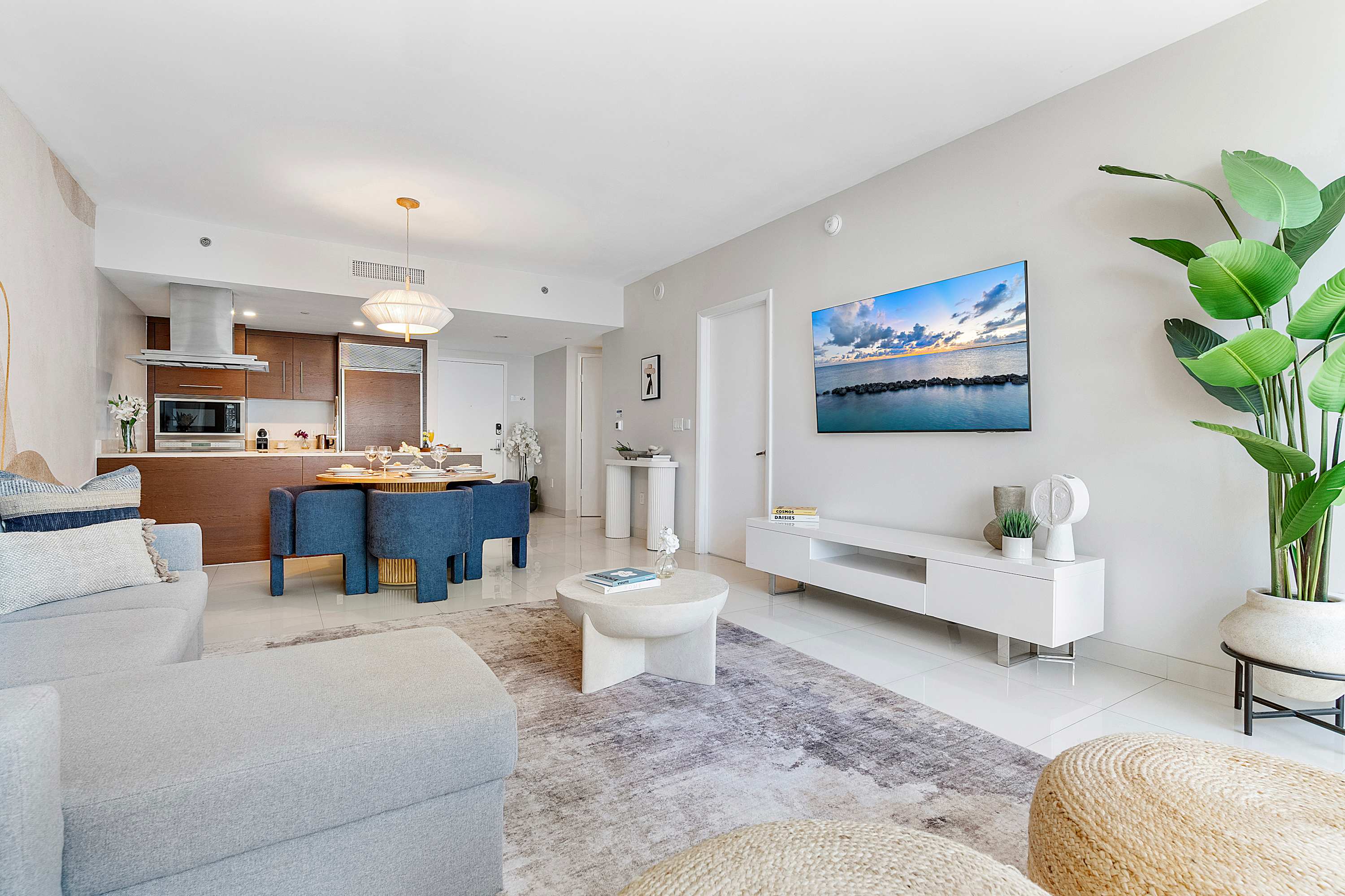 Open-concept living space with a grey sectional sofa, a round coffee table, and a wall-mounted TV, seamlessly flowing into a modern kitchen and dining area with white tile flooring throughout.