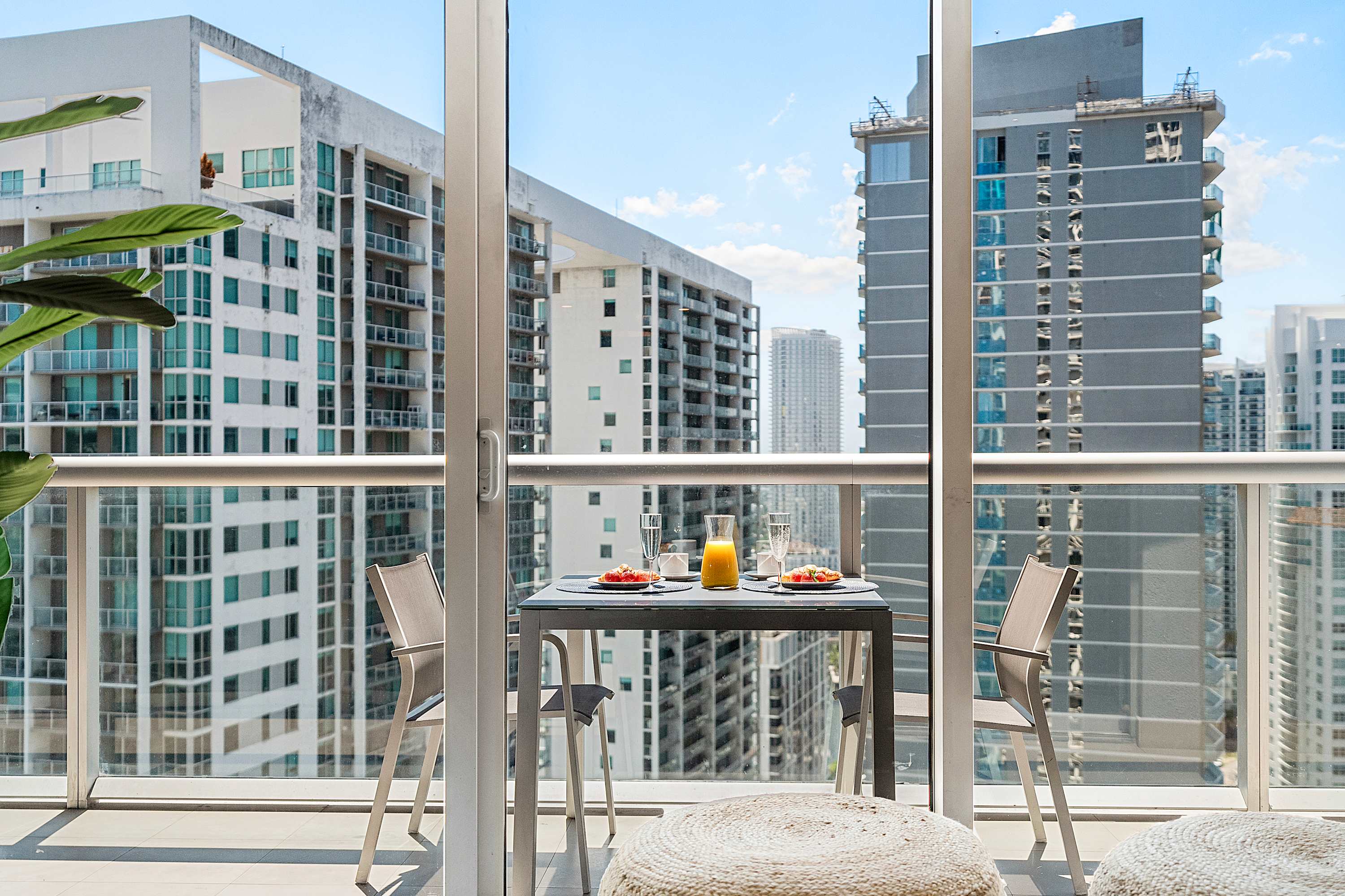 Outdoor balcony with a small dining table and two chairs, set against a backdrop of high-rise city buildings. The table is set with breakfast items, offering a scenic spot for morning meals.