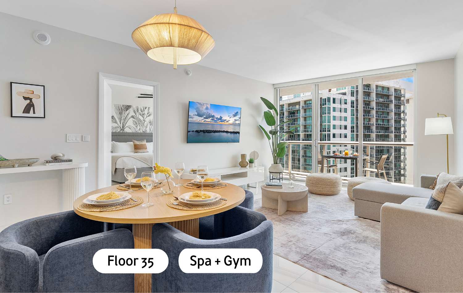 Modern open-concept living space featuring a light wood round dining table with four blue upholstered chairs, a large pendant light, and a living area with a TV and floor-to-ceiling windows offering city views.