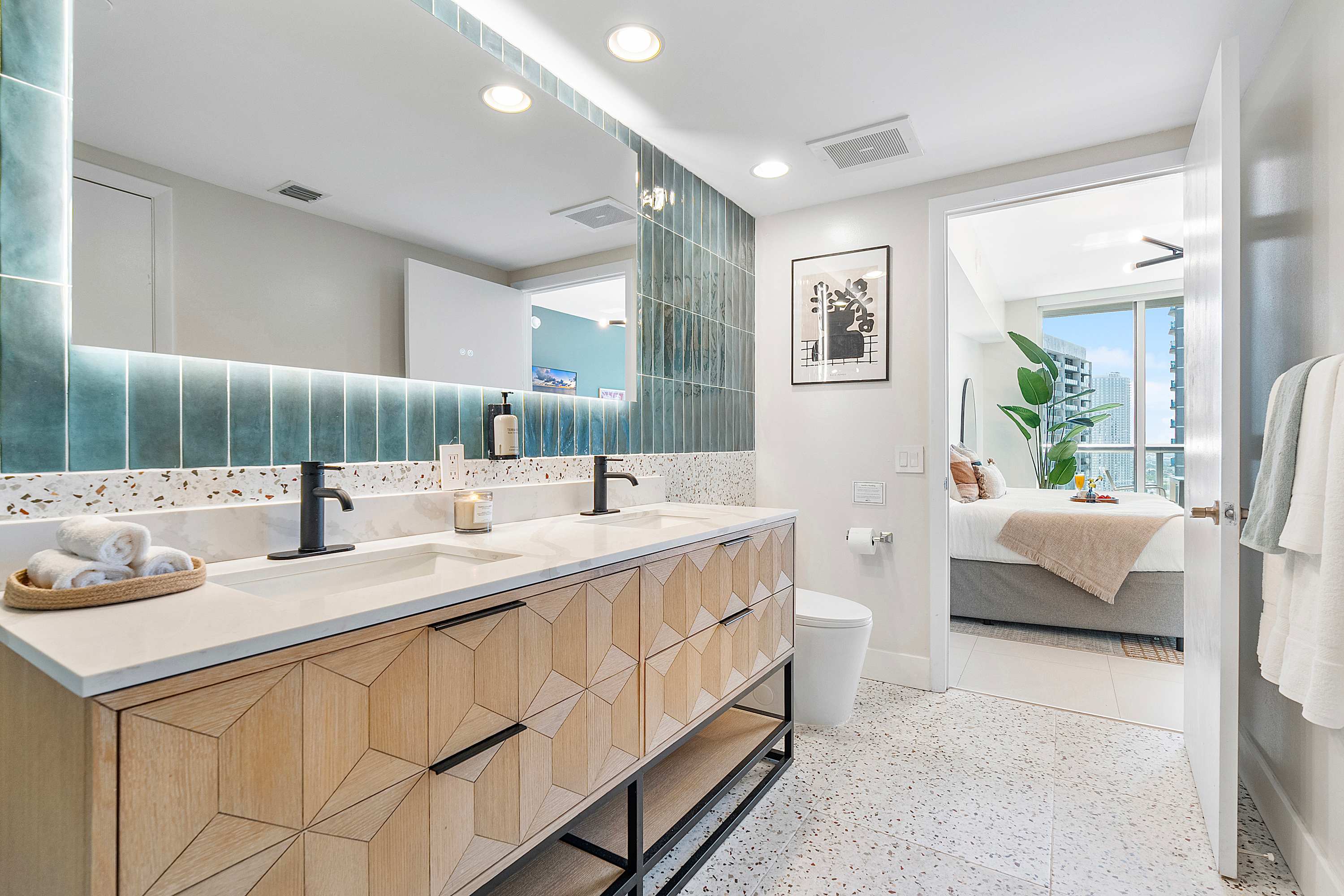 Modern bathroom with a double vanity featuring light wood cabinets, white marble countertops, and black fixtures. The wall behind the mirror is adorned with vertical teal tiles and a speckled backsplash.