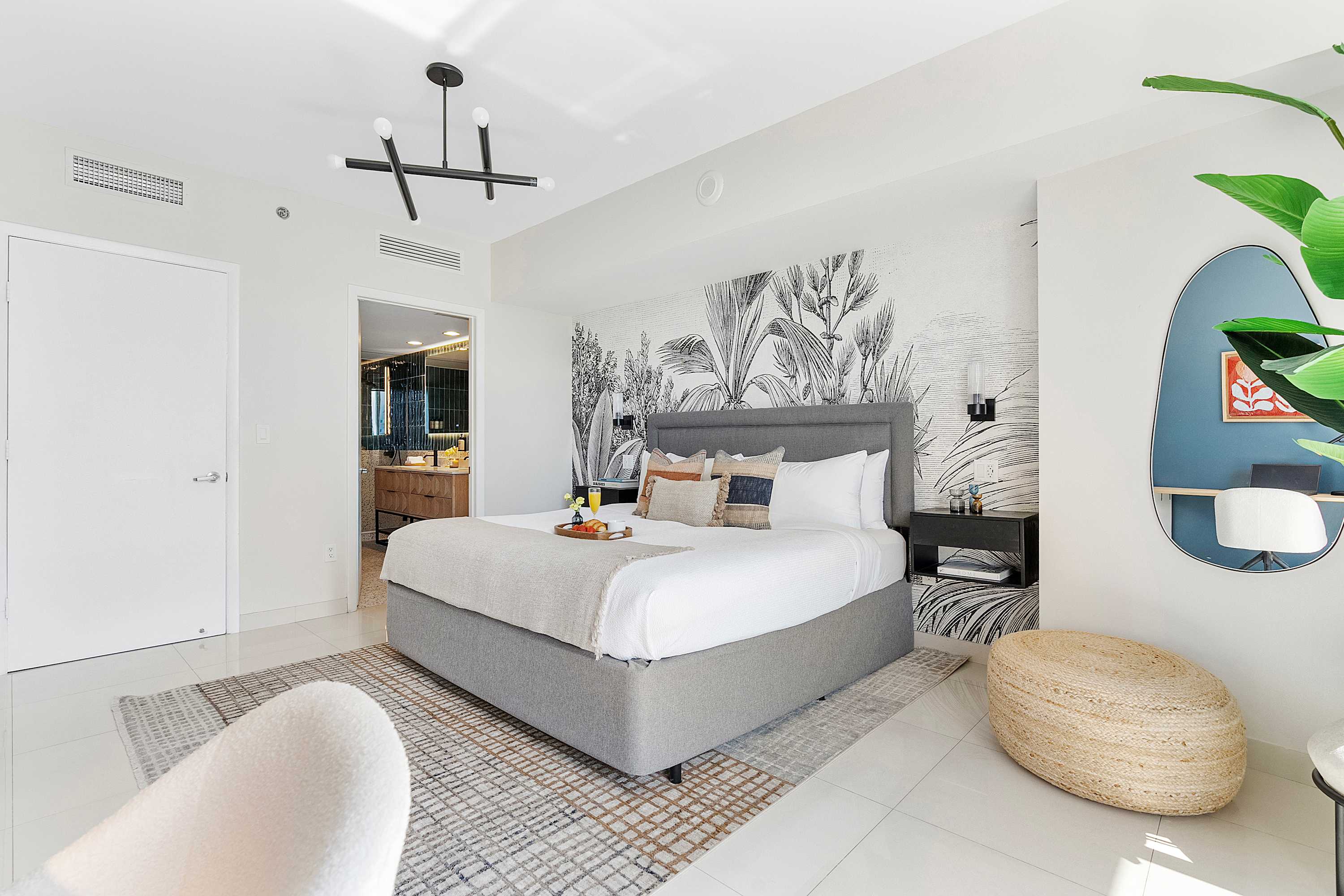Spacious bedroom featuring a gray upholstered king bed, a black nightstand, and a large plant. A doorway leads to a modern bathroom, and a woven pouf sits on the white tiled floor.