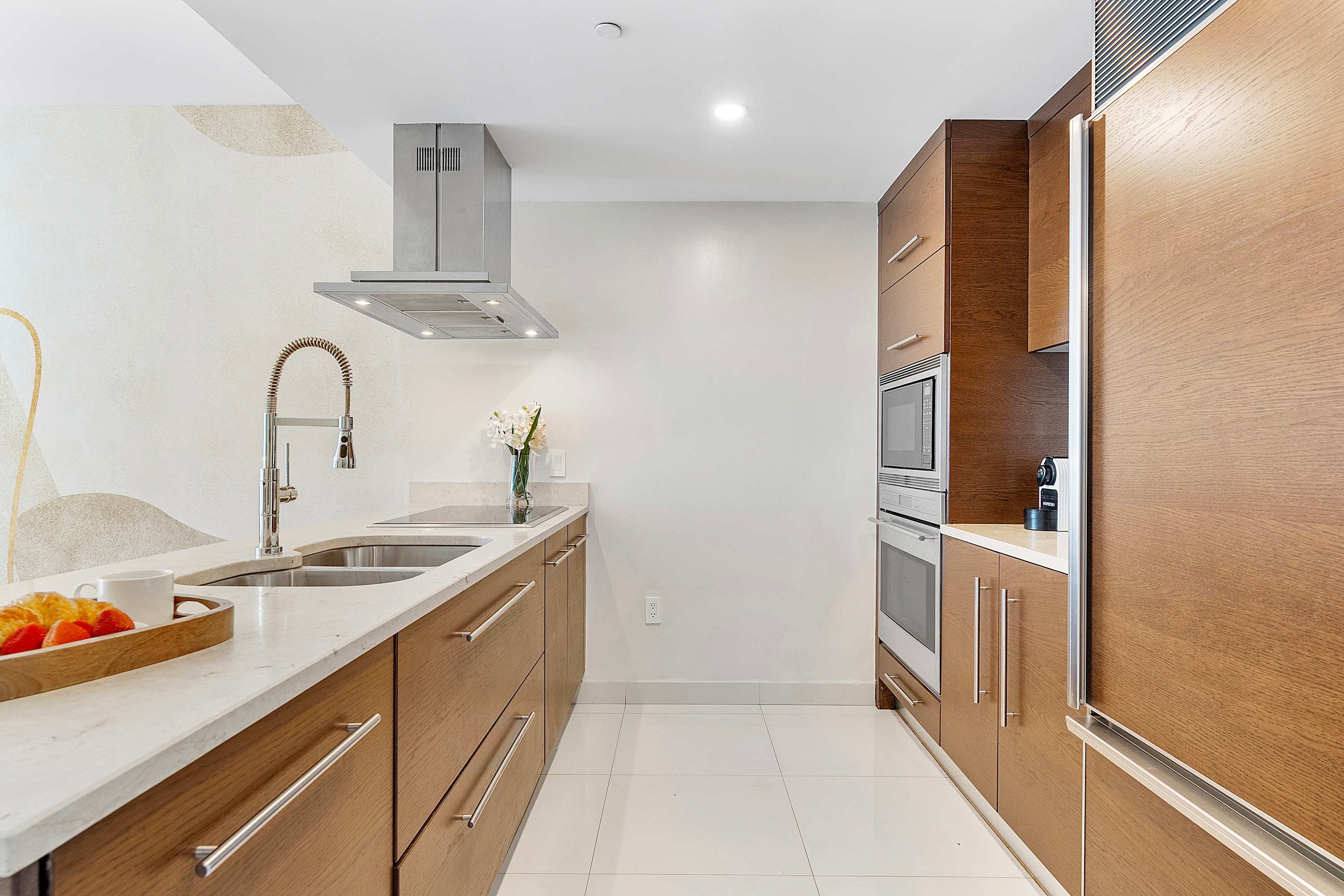 Modern galley kitchen featuring light wood cabinetry, white quartz countertops, and stainless steel appliances including a range hood and double sink. The floor is white tile, and walls are light with subtle abstract patterns.