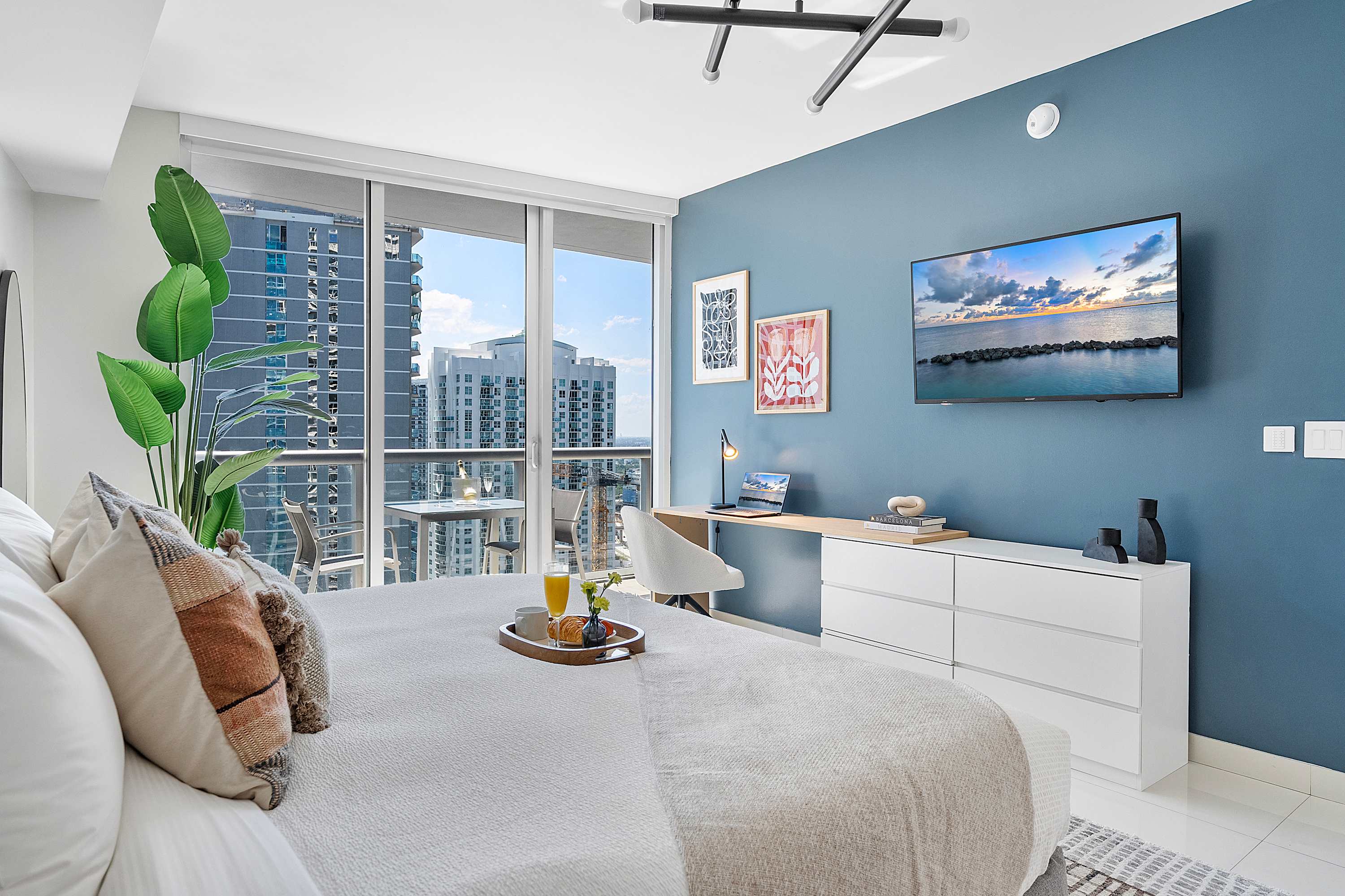 Bright bedroom with a large bed, a blue accent wall, a wall-mounted TV, and a desk area with a laptop, all illuminated by natural light from floor-to-ceiling windows leading to a balcony.