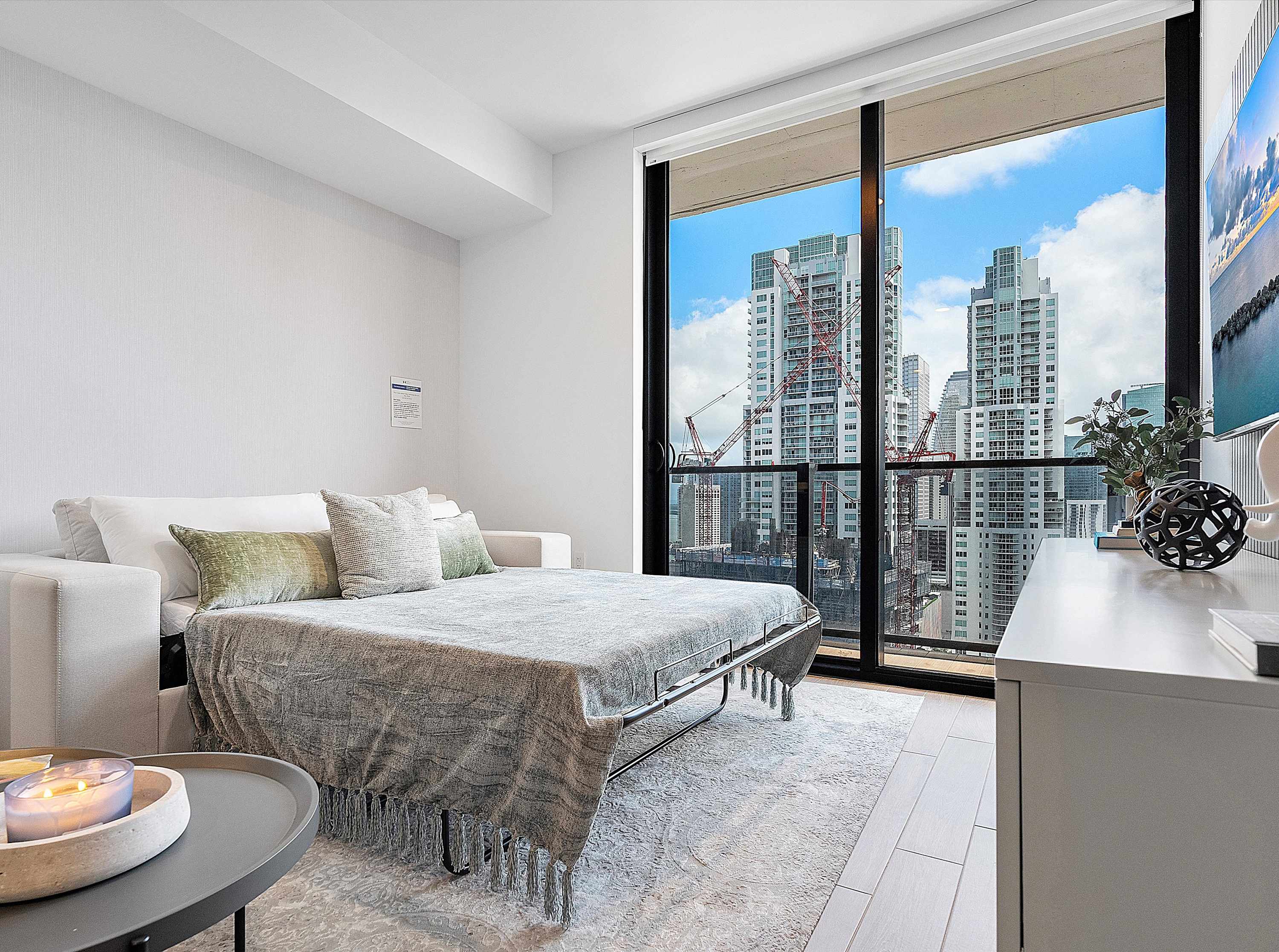 A bright living space featuring a sofa bed pulled out, covered with a grey throw, facing large floor-to-ceiling windows that offer panoramic city views with towering buildings and construction cranes.