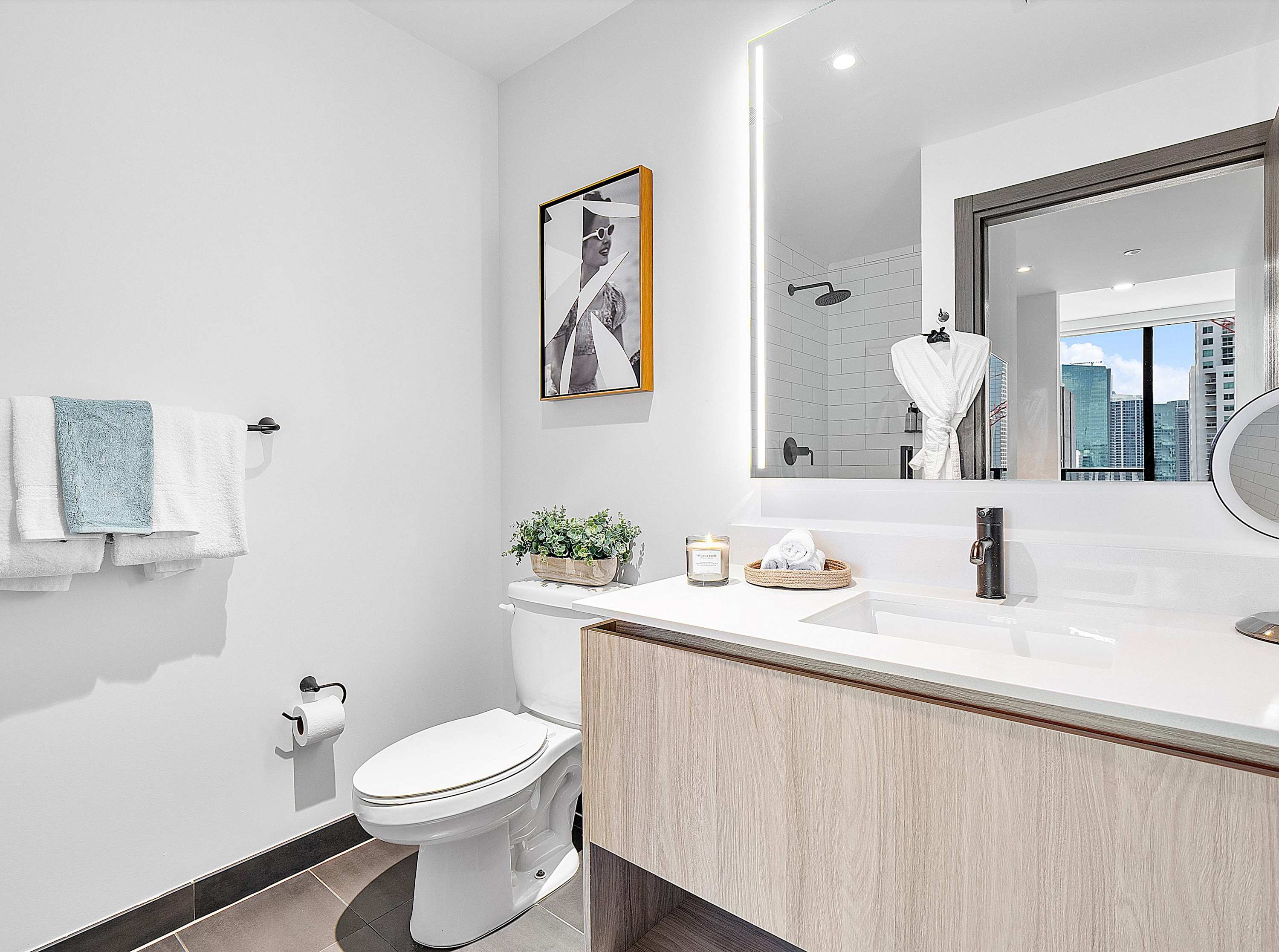 Bright, modern bathroom featuring a light wood double vanity with white countertops, a large illuminated mirror, and a white toilet. The shower, visible in the mirror, has white subway tiles and a city view reflection.