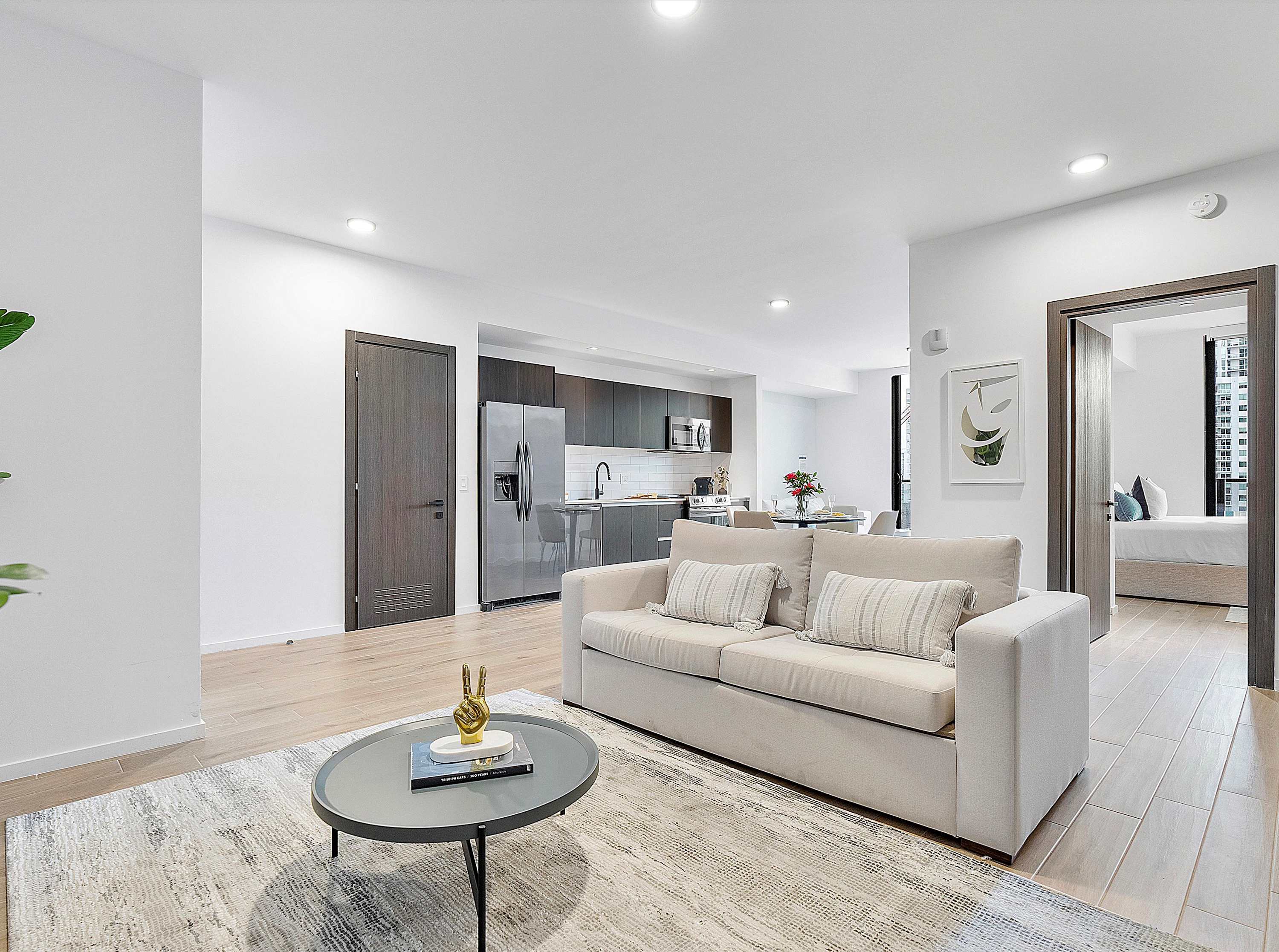 A contemporary living room with a light-colored sofa, a round grey coffee table, and a patterned rug, with an open view towards the modern kitchen and dining area in the background.