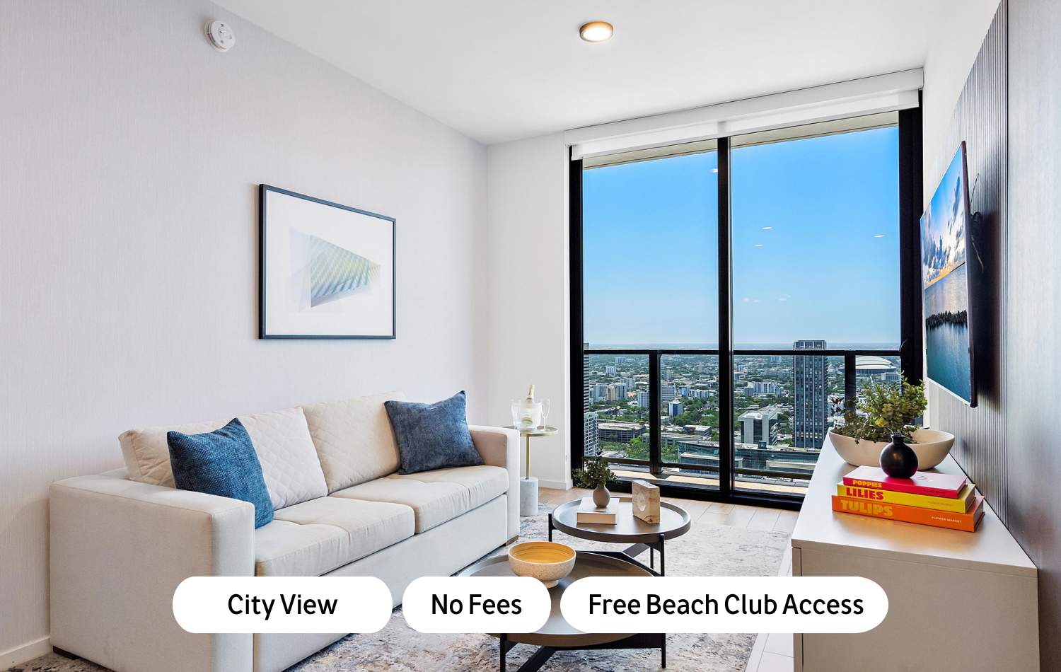 Modern living room featuring a light-colored sofa with blue accent pillows, two round coffee tables on a patterned rug, and a flat-screen TV. Floor-to-ceiling windows offer a panoramic city view.