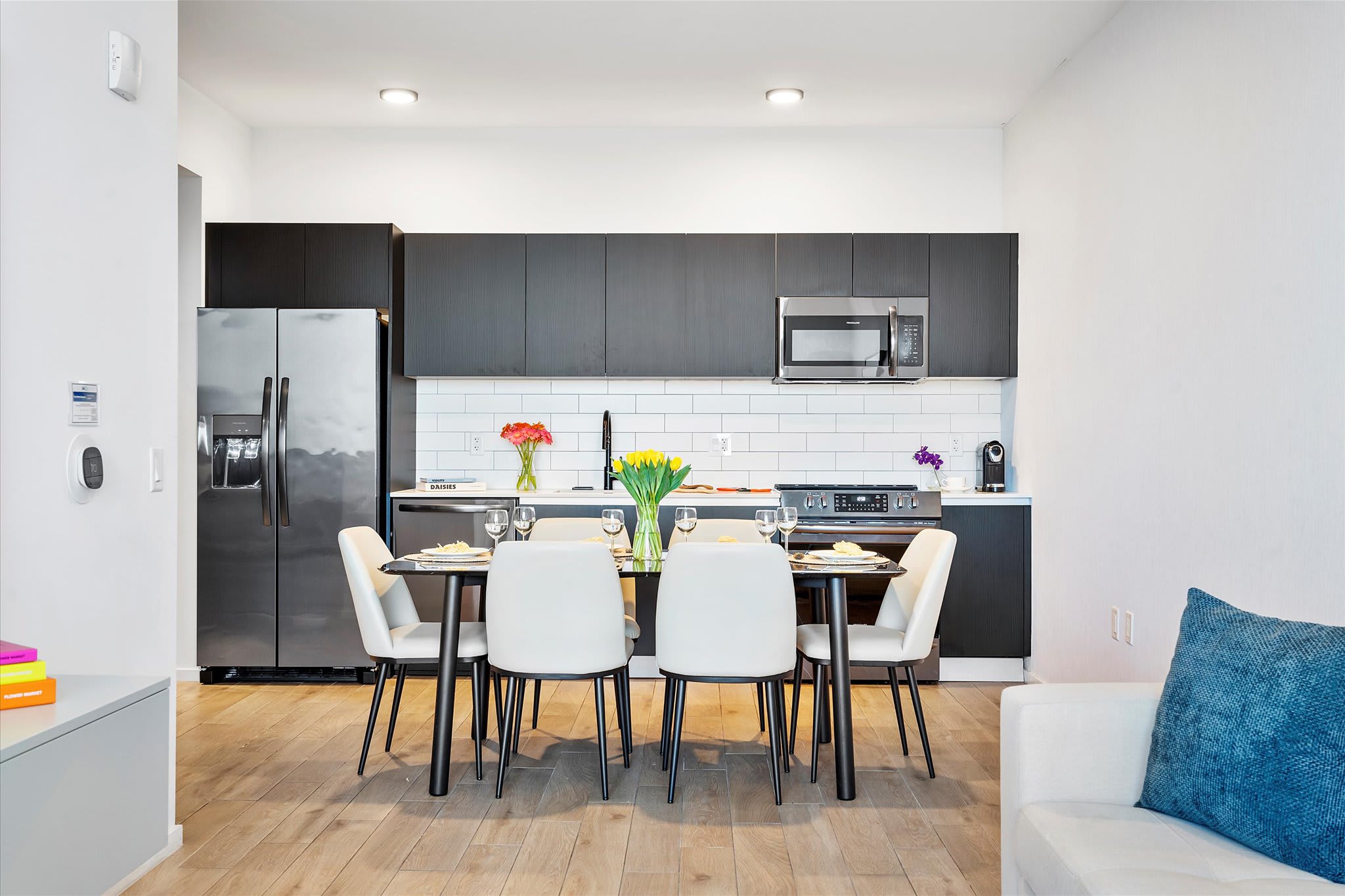 Modern kitchen featuring dark flat-panel cabinetry, white subway tile backsplash, and stainless steel appliances. A dining table with six white chairs and a floral centerpiece is centrally placed.