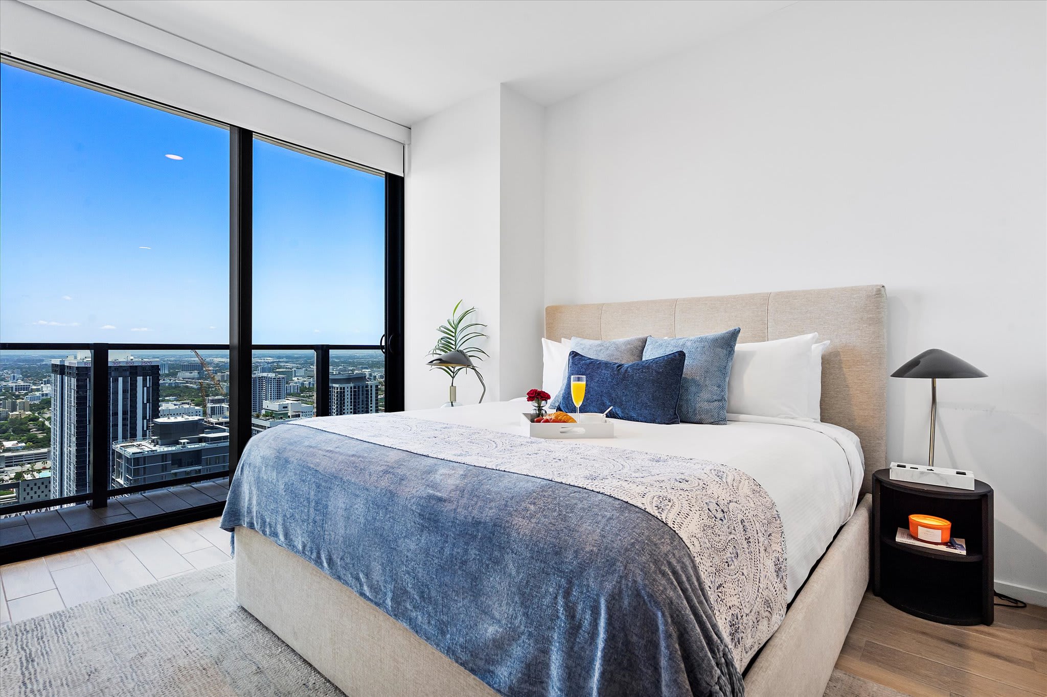 Bright bedroom with a queen-sized bed featuring a beige upholstered headboard and blue and white bedding. Floor-to-ceiling windows provide a stunning city view, complemented by a simple nightstand and lamp.