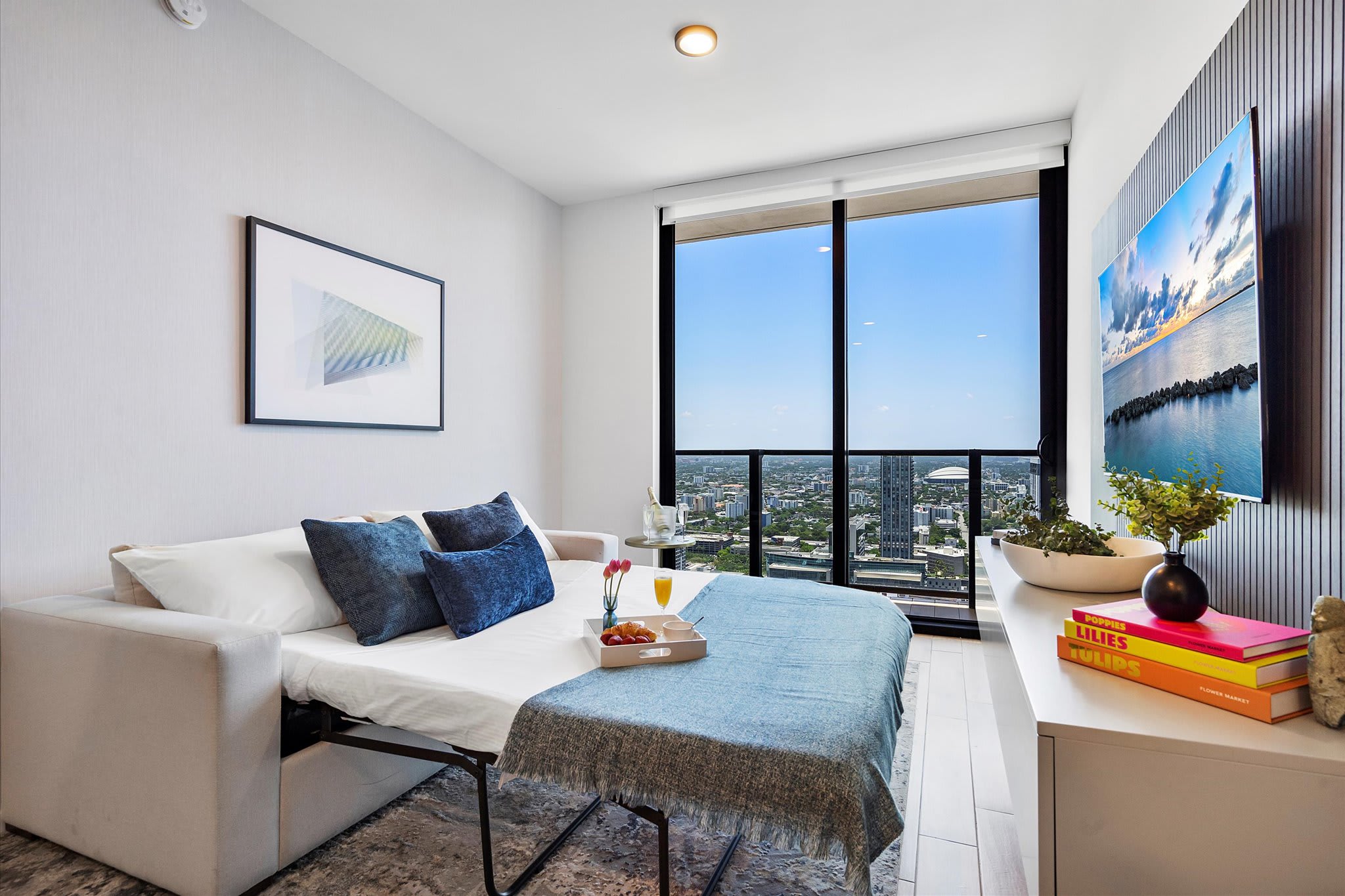 Living room configured as a secondary sleeping area with a pull-out sofa bed made up with blue bedding. Floor-to-ceiling windows offer a city view, and a flat-screen TV is mounted on the wall.
