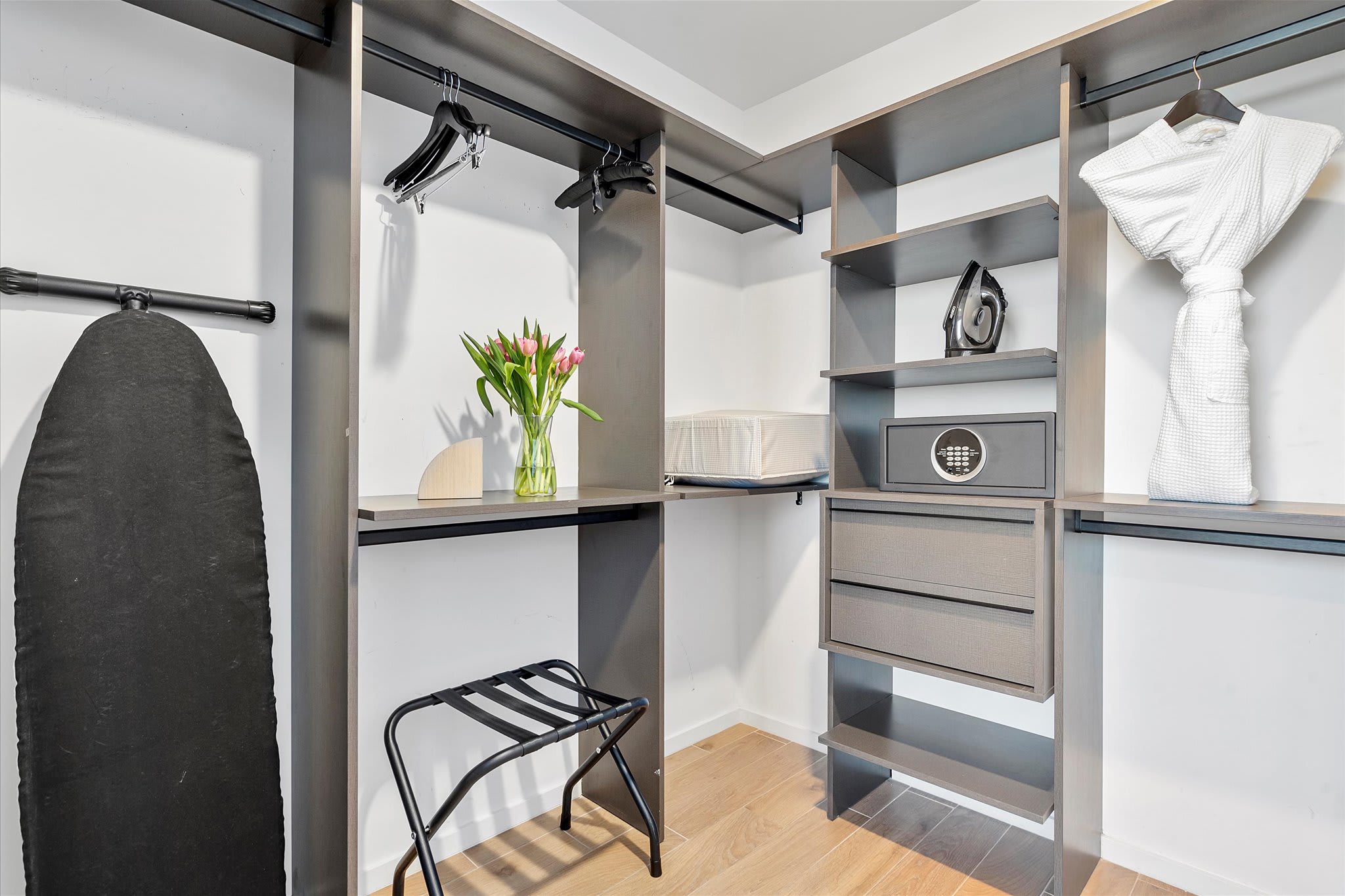 Walk-in closet with built-in dark shelving and hanging rods, equipped with an ironing board, a luggage rack, and a small safe. A vase with pink tulips adds a decorative touch.