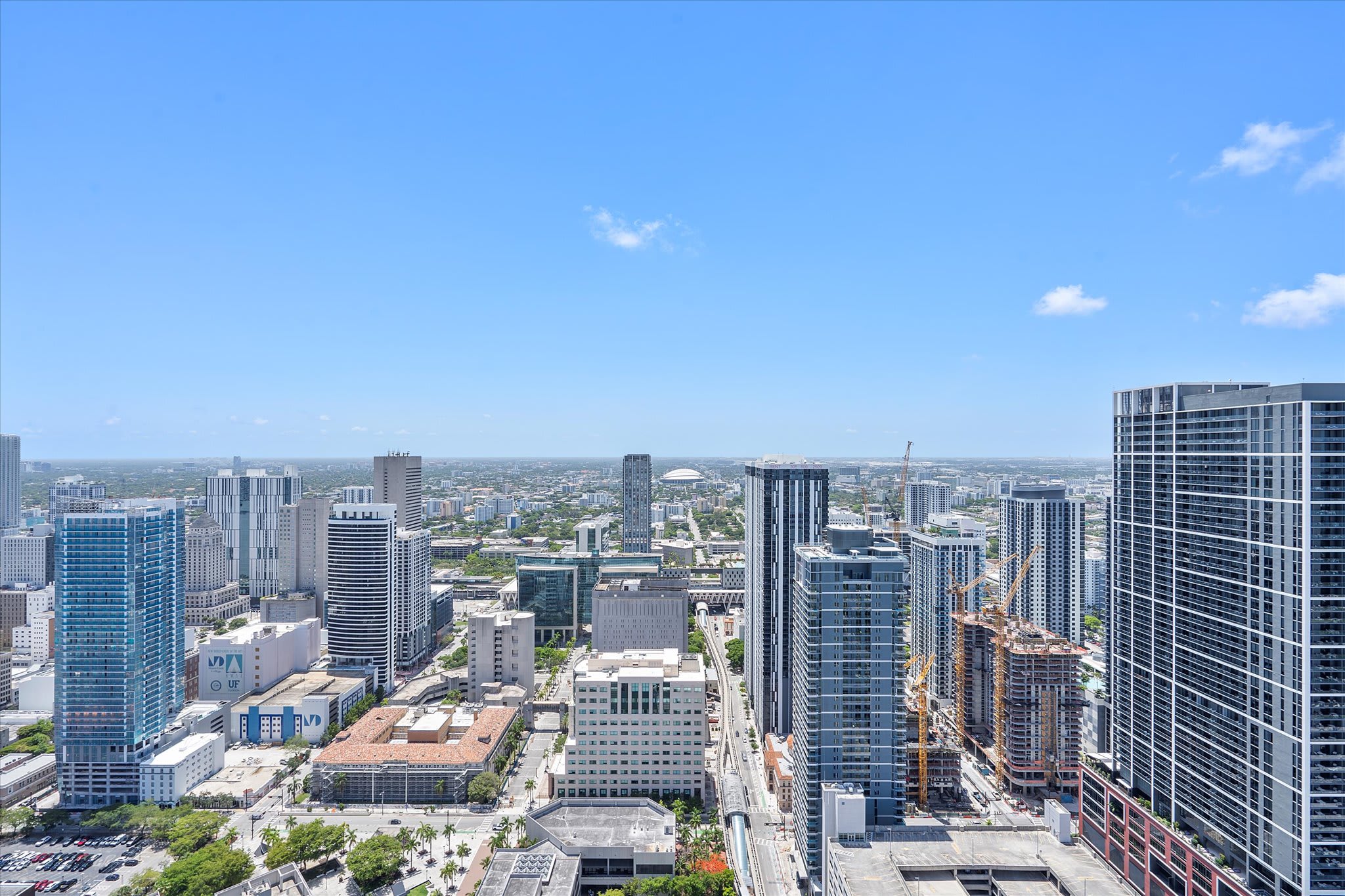 A high-angle aerial view captures a sprawling urban landscape under a clear blue sky, featuring numerous high-rise buildings, a stadium in the distance, and a visible elevated train track winding through the city.