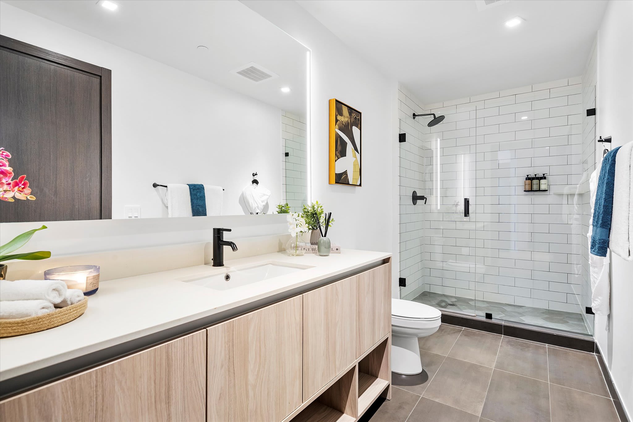 Contemporary bathroom with a large vanity featuring light wood cabinets, a white countertop, and a rectangular mirror with integrated lighting. A spacious walk-in shower with a glass enclosure and white subway tiles is visible.