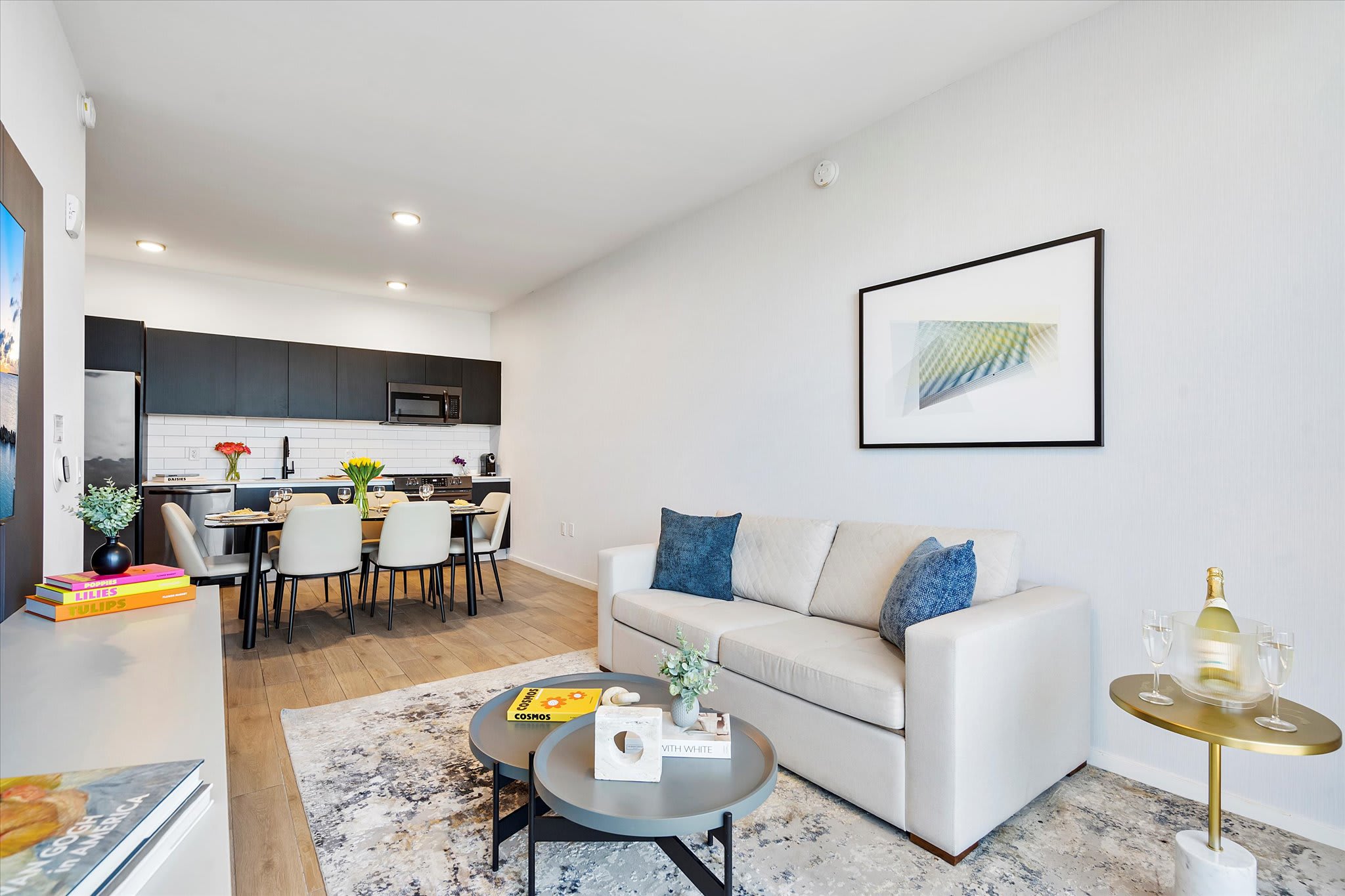 Open-concept living area with light wood flooring, featuring a cream-colored sofa, two round coffee tables, and a patterned rug. The modern kitchen and dining area are visible in the background.