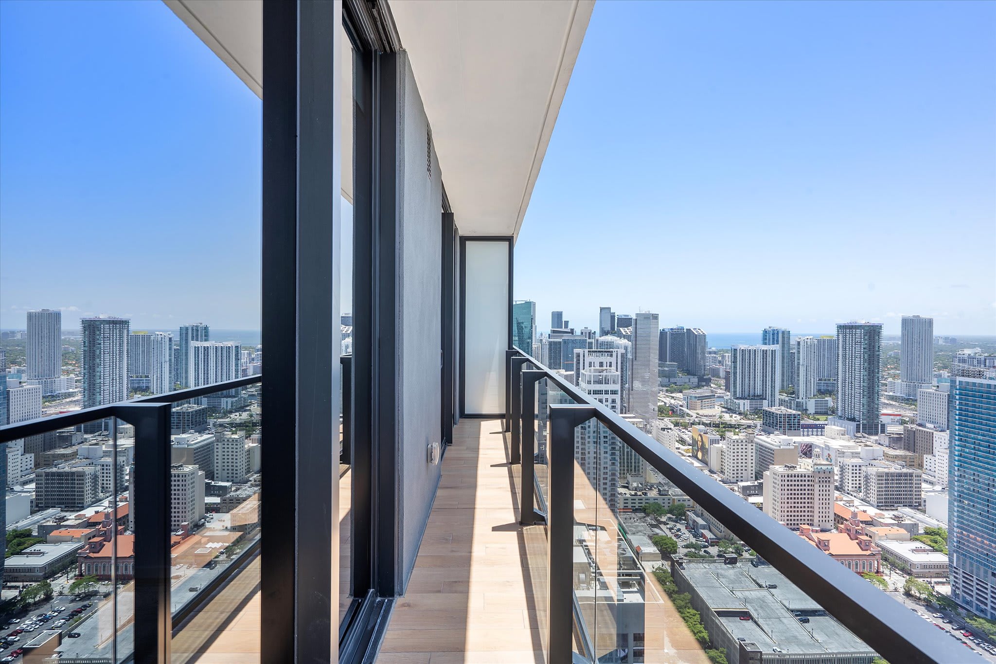 A long, narrow private balcony with a glass railing, offering an expansive view of a bustling city skyline with numerous high-rise buildings under a clear blue sky. The balcony floor is light wood.