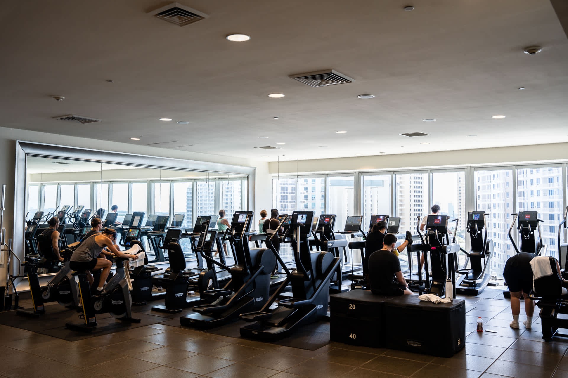 A modern, spacious gym with multiple cardio machines, including treadmills, ellipticals, and stationary bikes, where people are actively exercising. Large windows offer city views, complemented by a mirrored wall reflecting the equipment and activity.