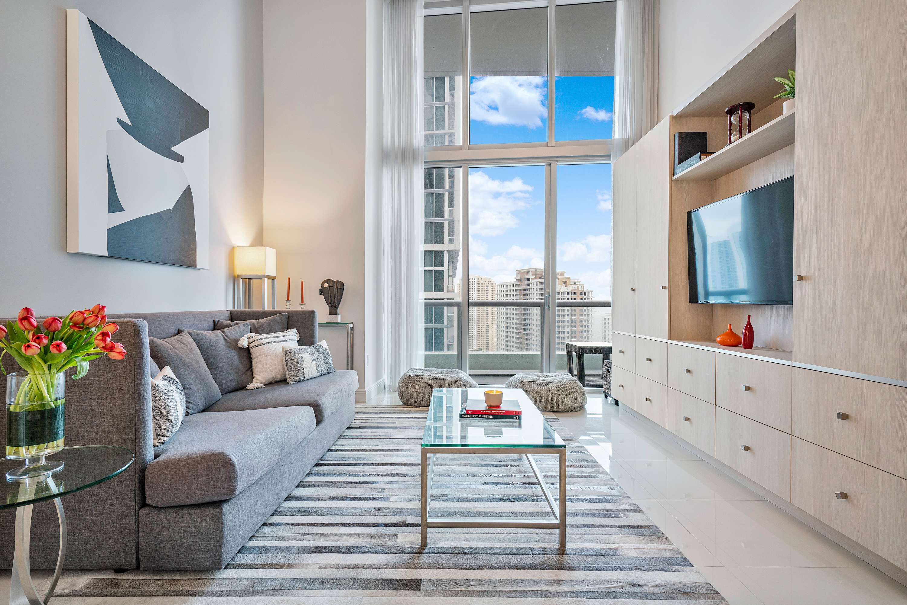Bright living room with a long gray sofa, striped area rug, and floor-to-ceiling windows showcasing city views. A wall-mounted TV and light wood built-in storage complete the modern aesthetic.