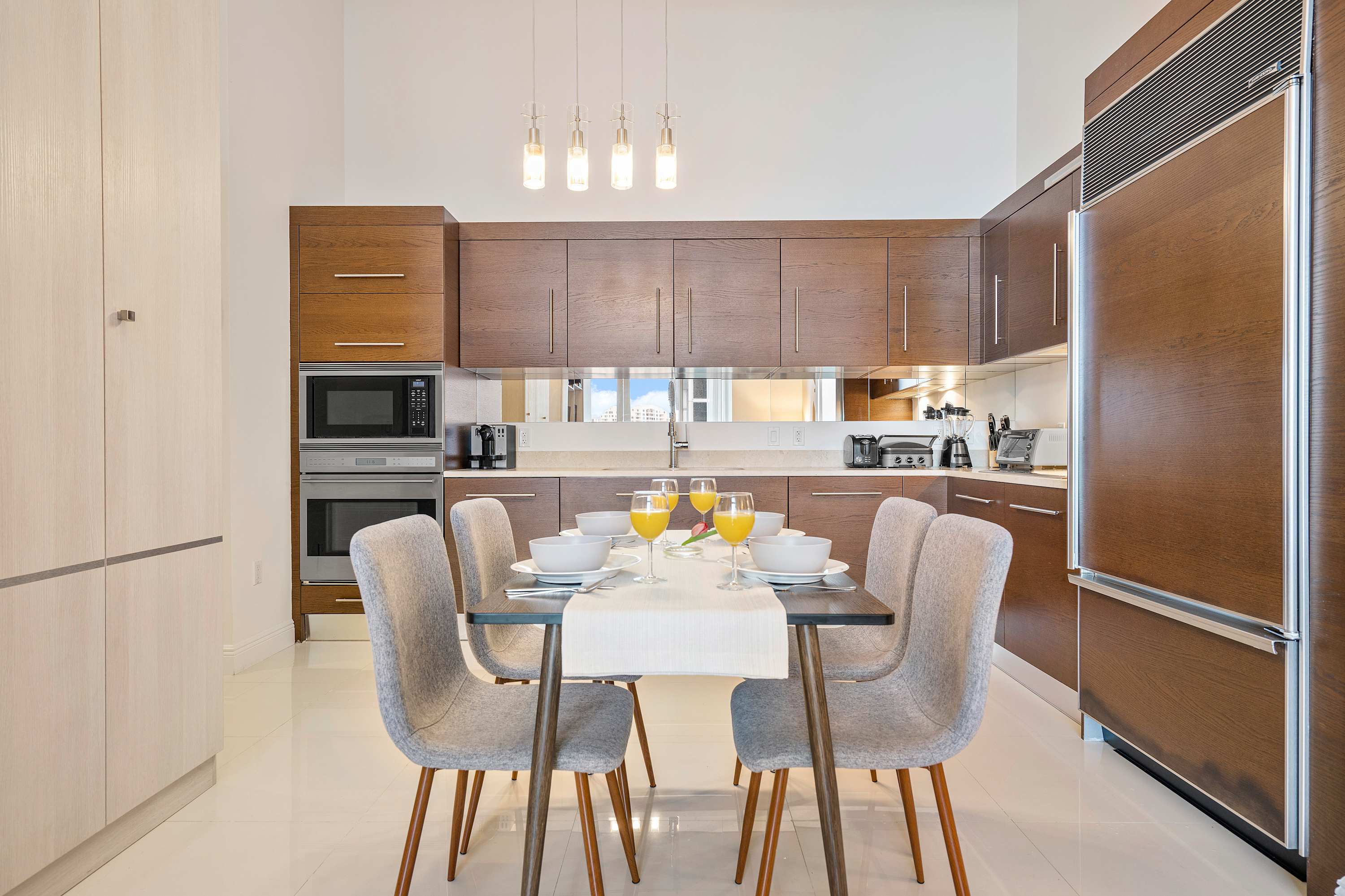 Contemporary kitchen and dining area featuring rich wood cabinetry, integrated refrigerator, and a white countertop. A dining table with four upholstered chairs is neatly set, under stylish pendant lighting.