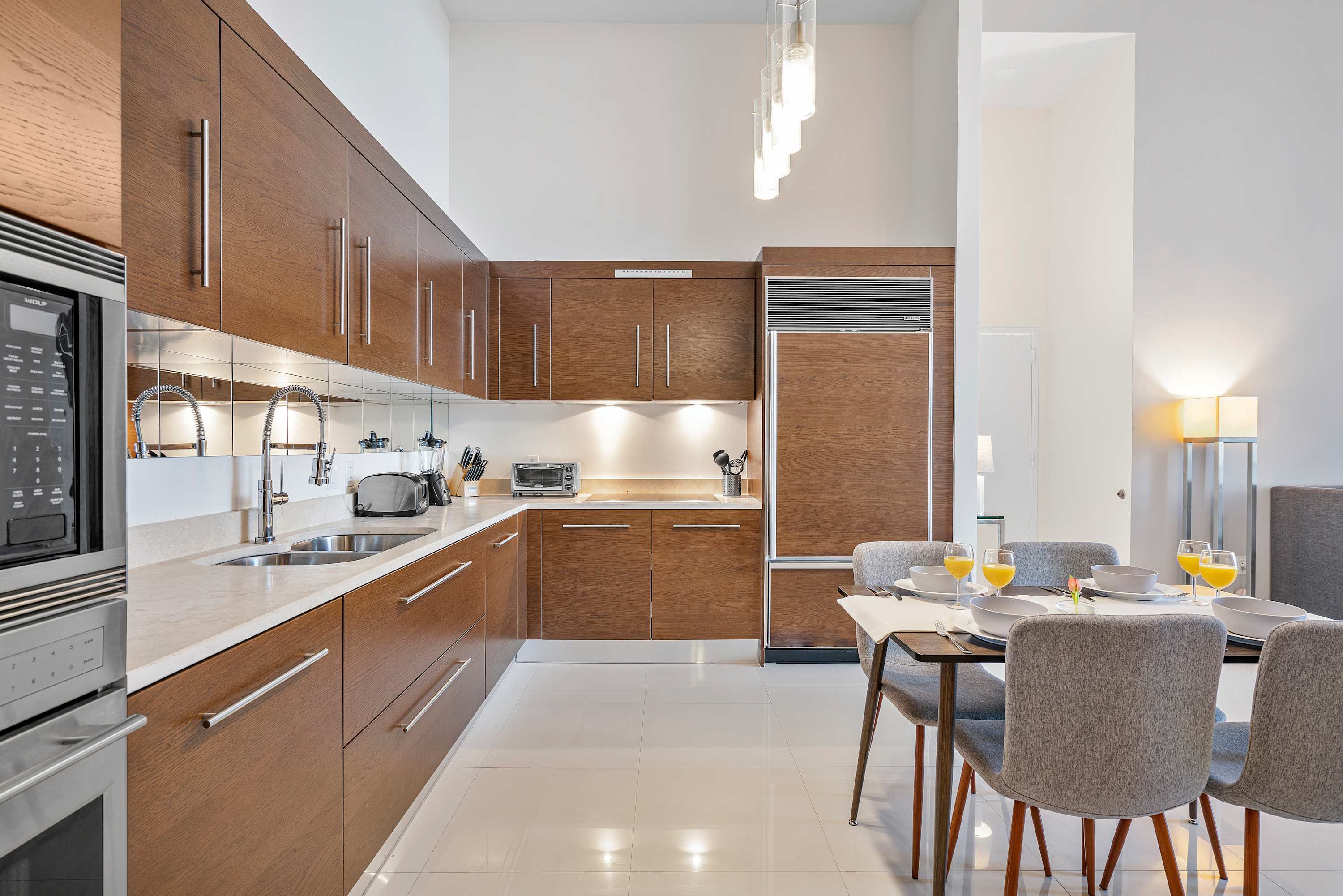 Sleek, modern kitchen with dark wood cabinetry, stainless steel appliances, and a mirrored backsplash. A dining table with four chairs is set for a meal, illuminated by contemporary pendant lights.