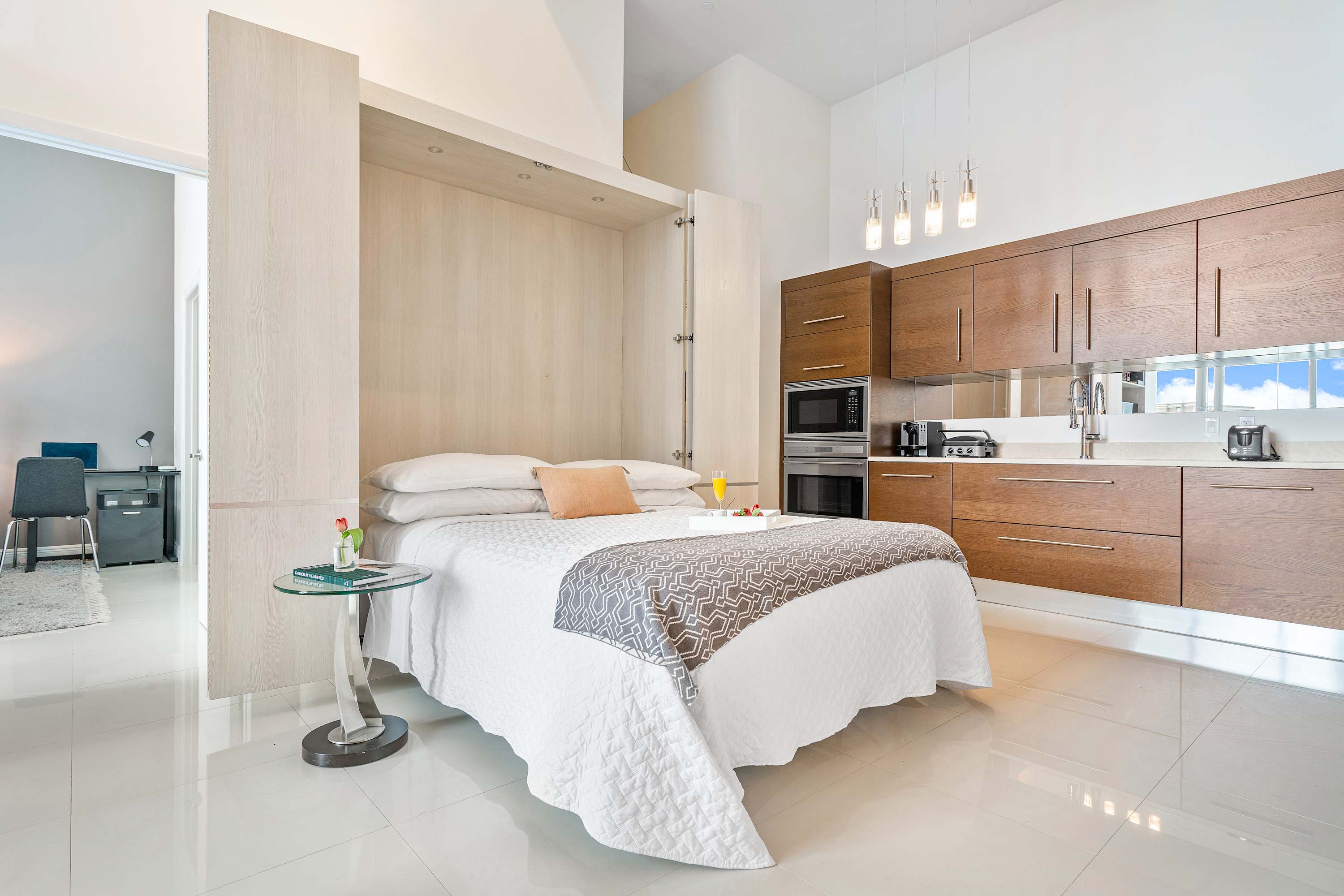 Open-concept living area featuring a pull-down Murphy bed with white bedding, integrated into light wood cabinetry. A modern kitchen with dark wood cabinets is visible in the background, all under high ceilings.