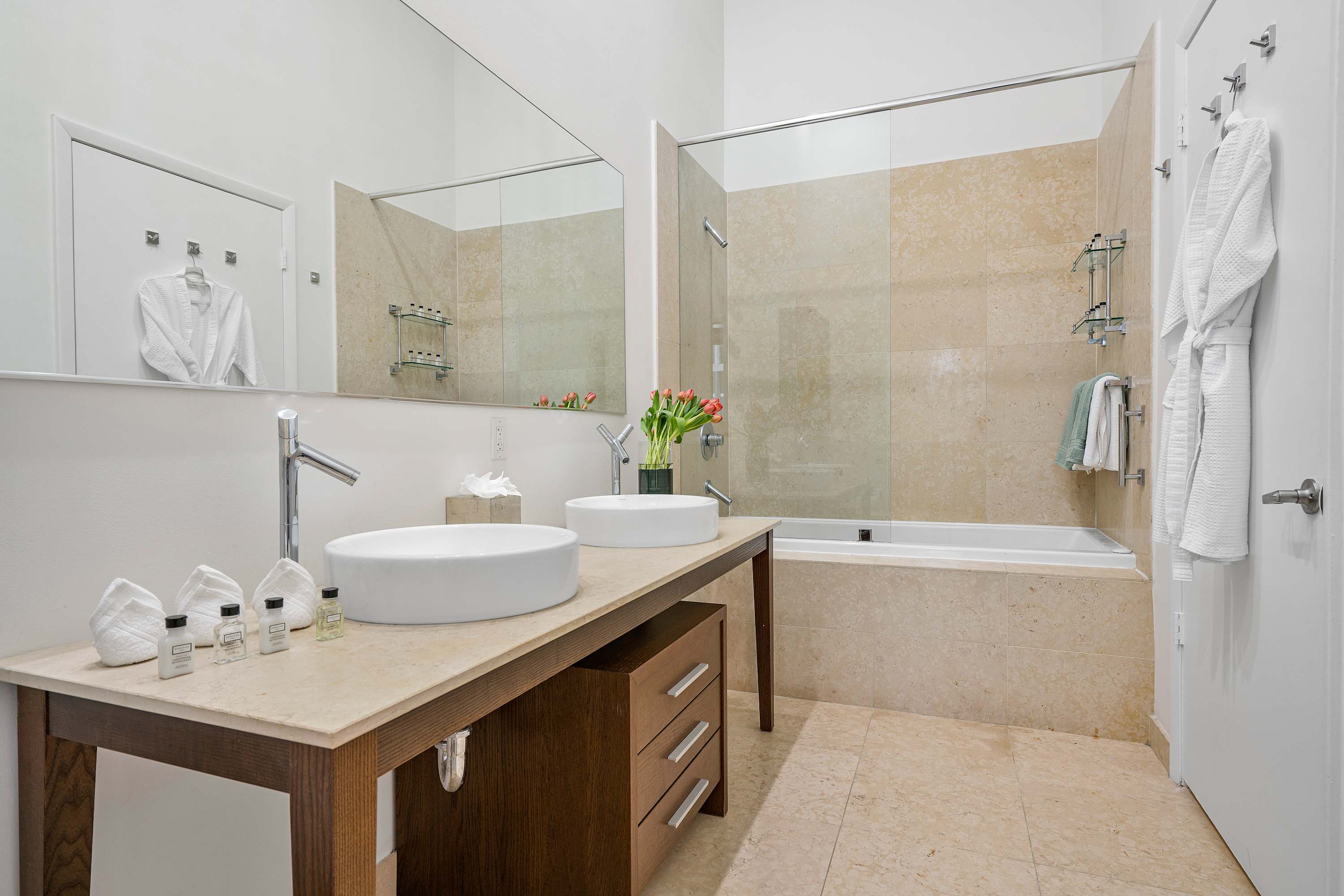 Contemporary bathroom with a double vanity, vessel sinks, a large mirror, and a tiled alcove bathtub with a glass shower screen, featuring white bathrobes hanging on the door.