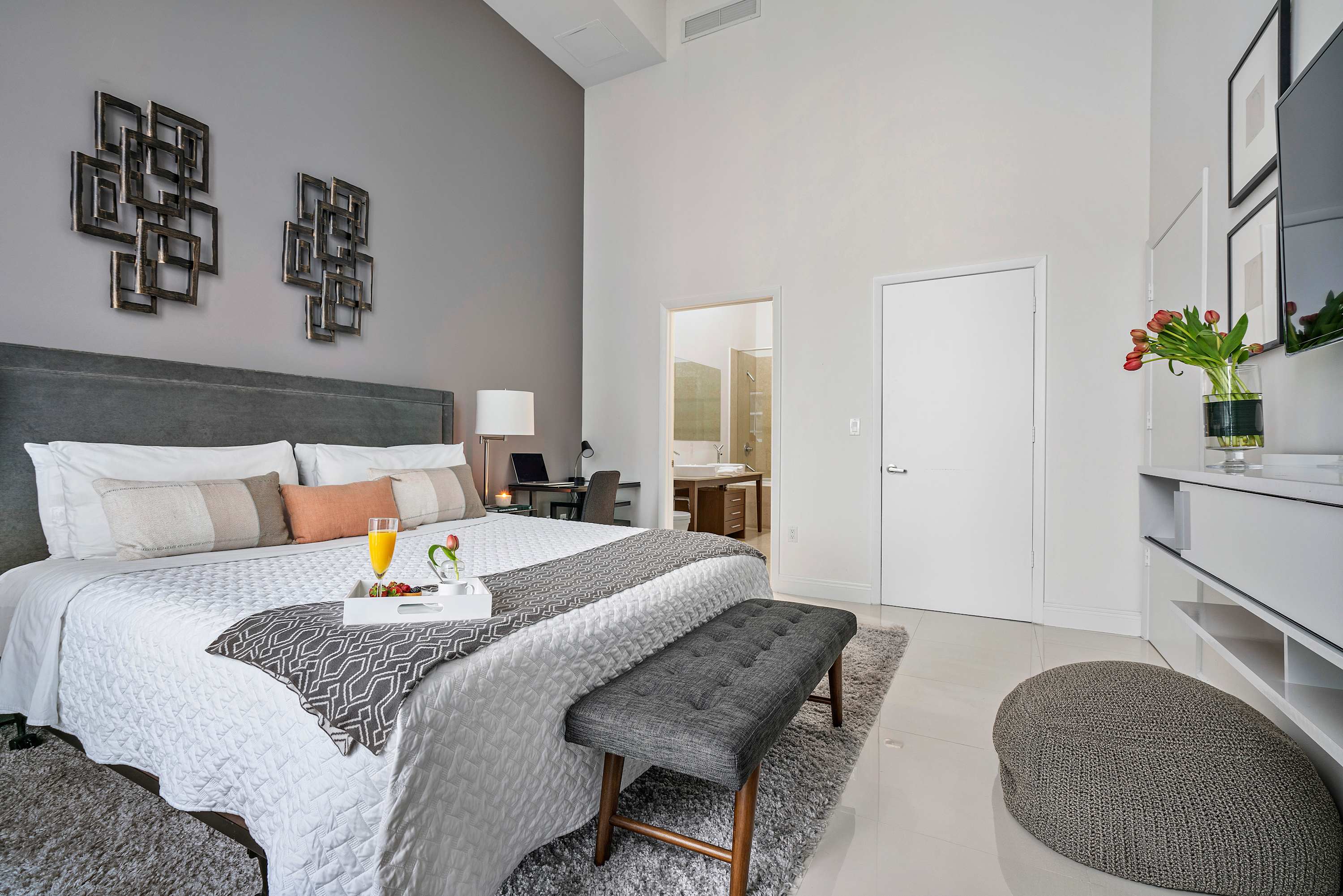 Elegant bedroom with a large bed, gray upholstered headboard, and a plush area rug. A small desk is visible, and an open doorway leads to an en-suite bathroom, all under high ceilings.