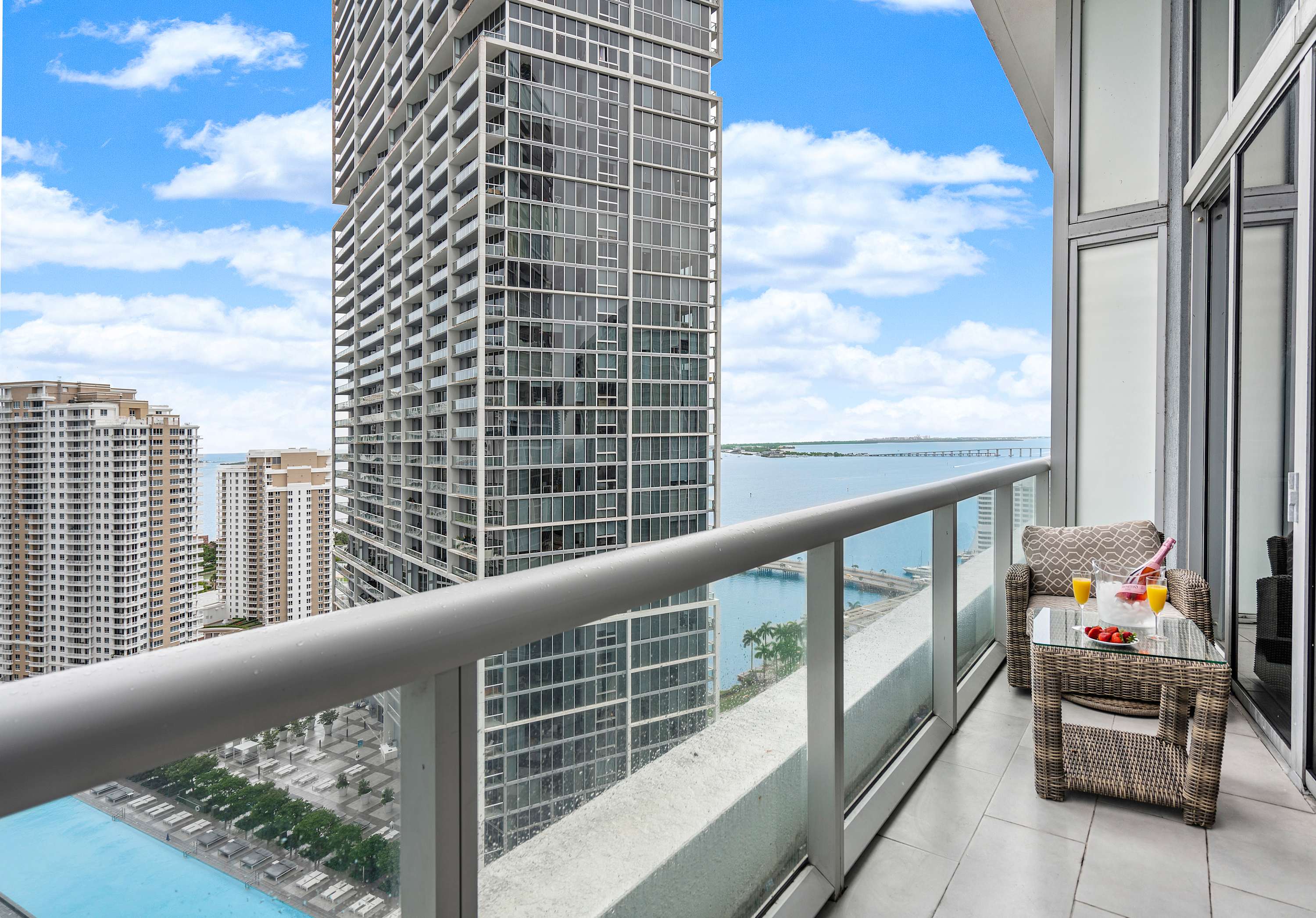 Modern high-rise balcony with a sleek glass railing, featuring a small wicker table set with refreshing drinks and fresh fruit, offering panoramic views of the city skyline, bay, and a distant bridge under a bright blue sky.