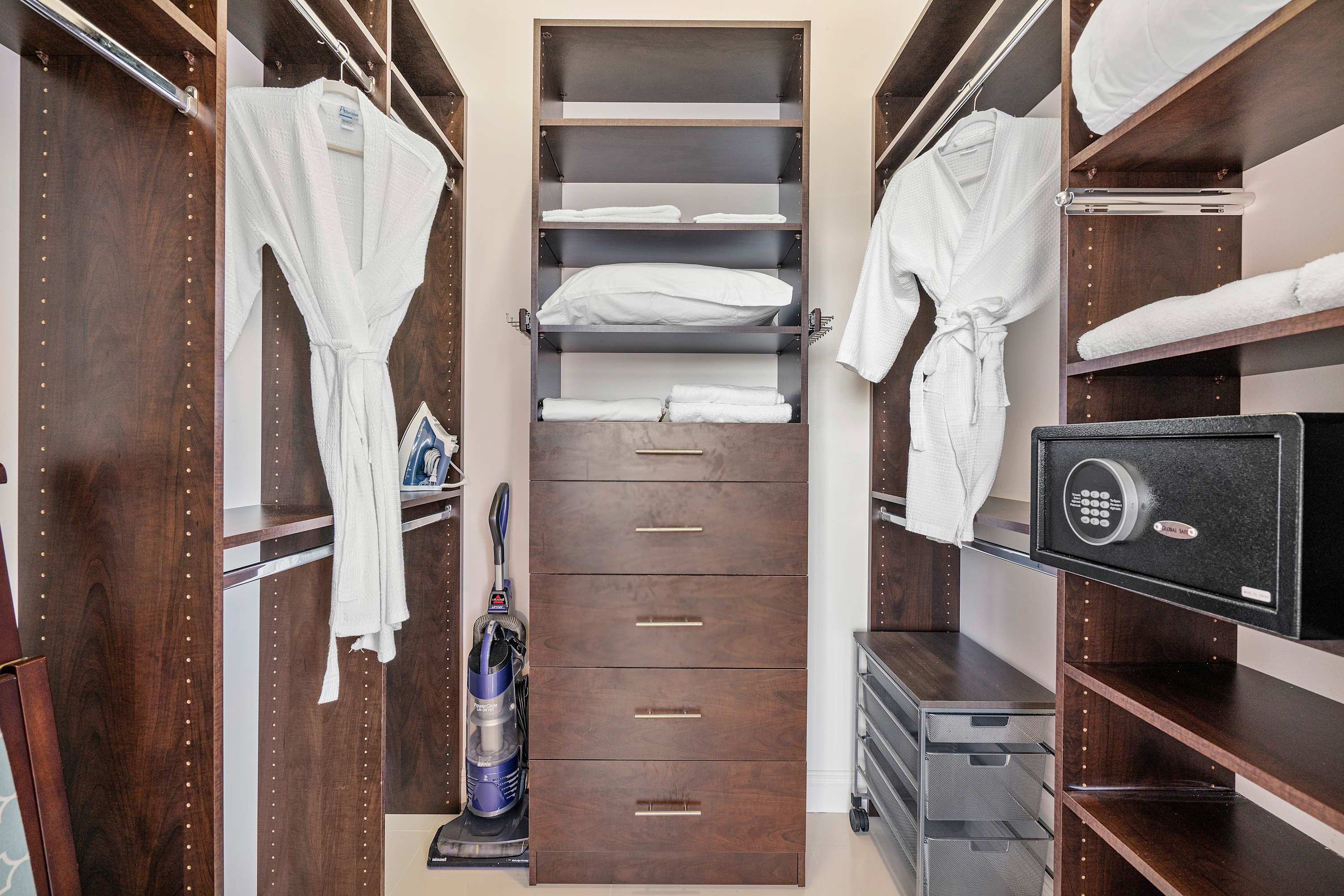 Organized walk-in closet with dark wood shelving and drawers, featuring hanging bathrobes, an iron, a vacuum cleaner, and a safe, providing ample storage space.