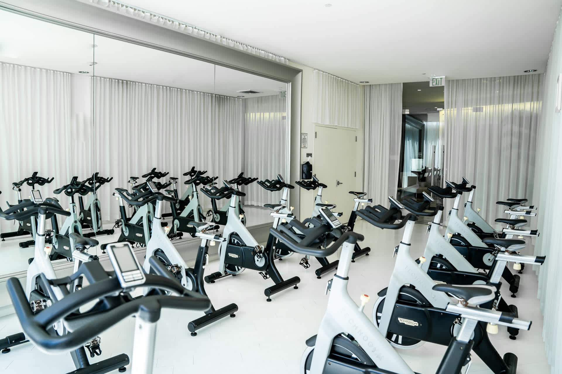 A bright, spacious fitness room equipped with numerous stationary spin bikes arranged in rows, featuring large mirrors along one wall and white curtains. The room appears ready for a group exercise class.