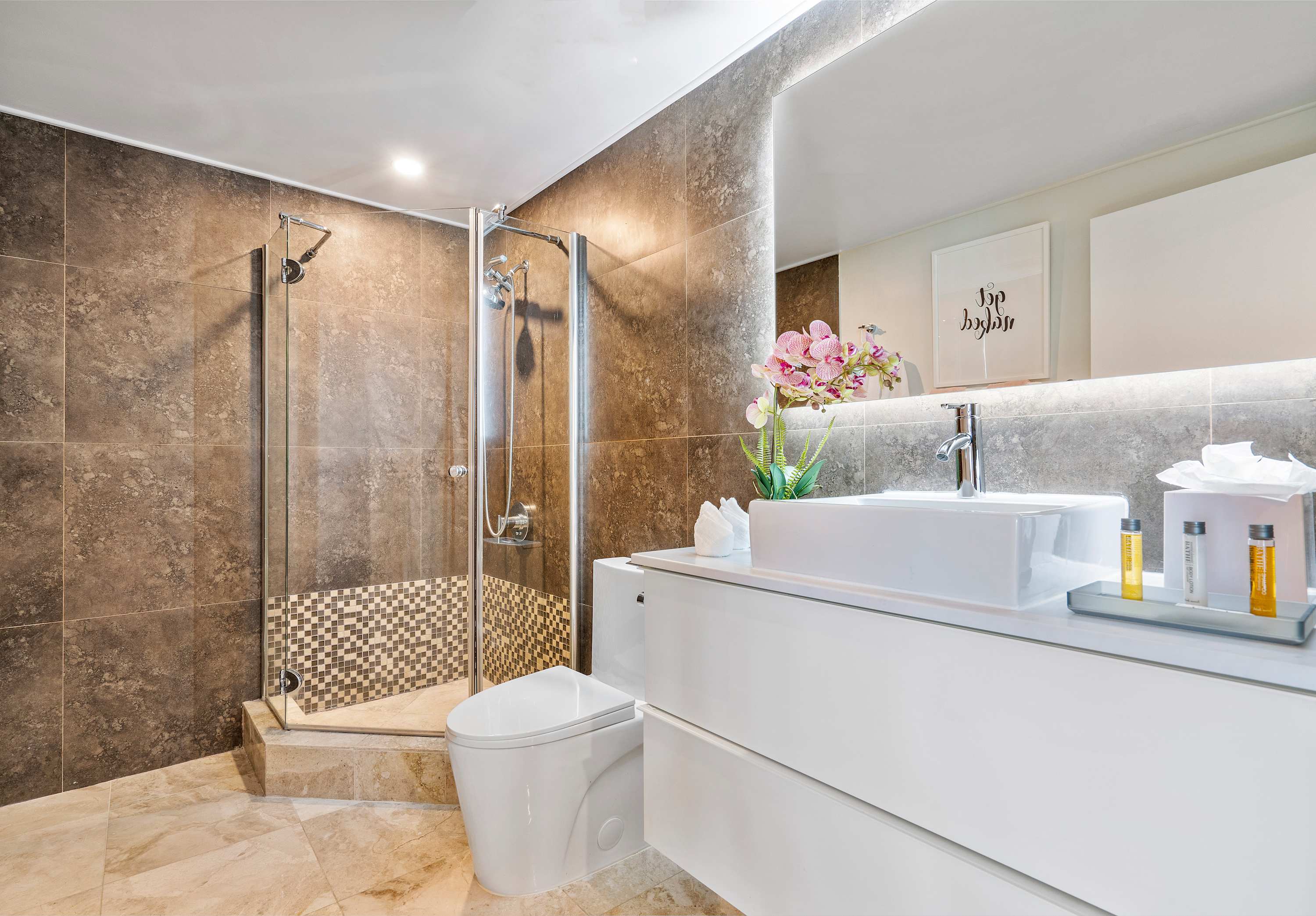 Modern bathroom with dark brown tiled walls, light marble floor, a glass-enclosed shower with mosaic accents, and a white floating vanity with a vessel sink and backlit mirror.