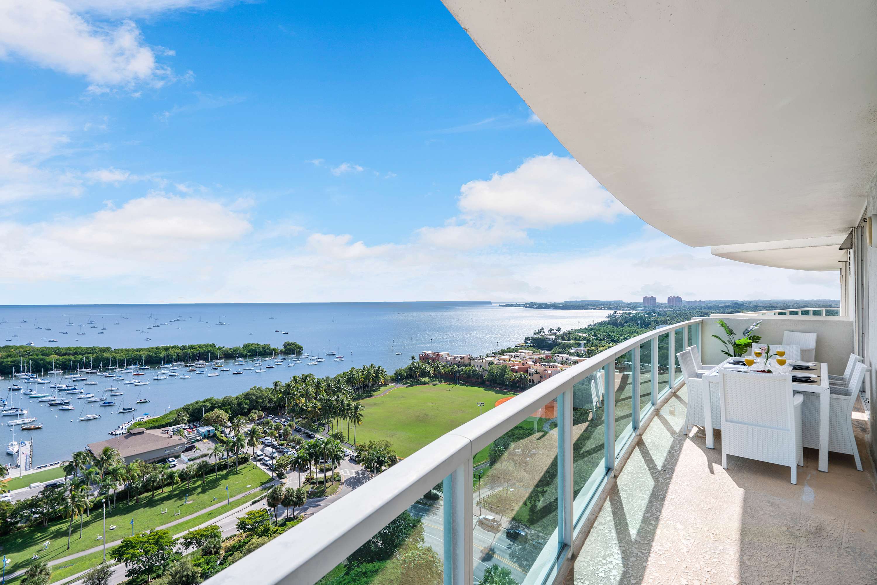 Expansive curved balcony with glass railings, offering panoramic views of a bay filled with boats, lush green areas, and distant cityscapes under a blue sky with white clouds. A white outdoor dining set is visible.