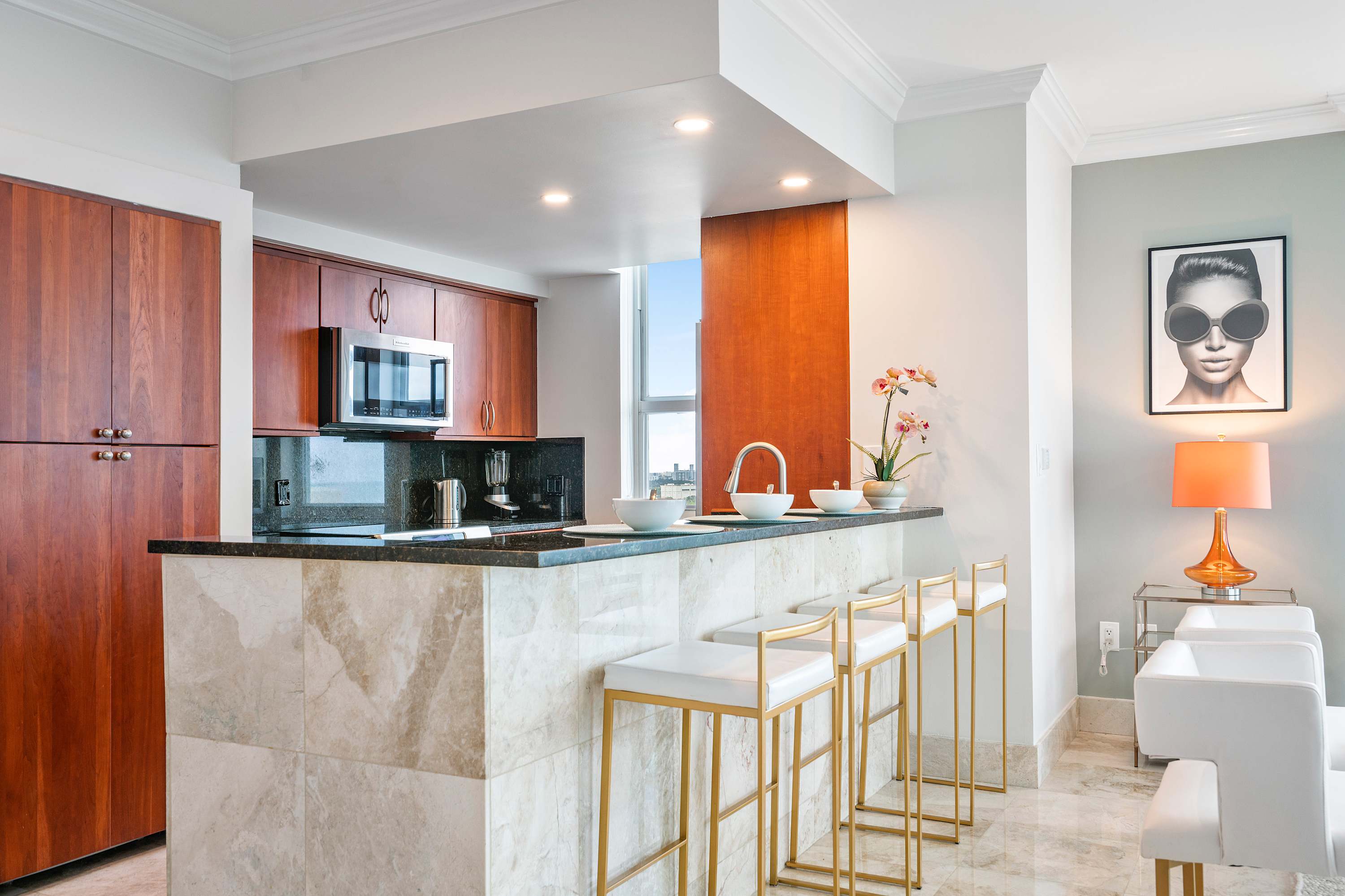 Modern kitchen with dark wood cabinetry, black granite countertops, stainless steel appliances, and a breakfast bar with gold-accented stools, overlooking the living area.