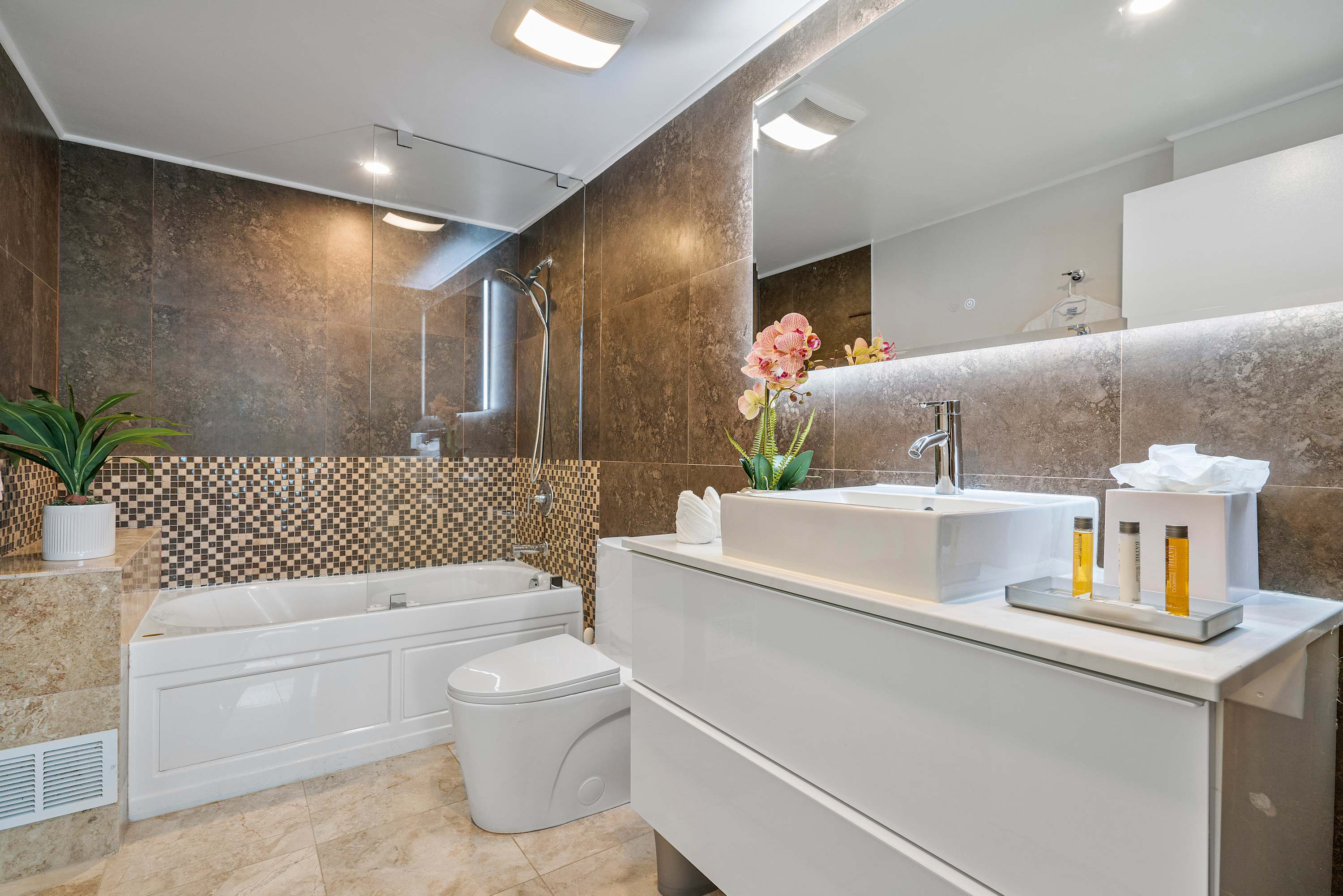 Modern bathroom with dark brown tiled walls, a white floating vanity with a vessel sink, a toilet, and a bathtub with a shower, illuminated by overhead lighting.