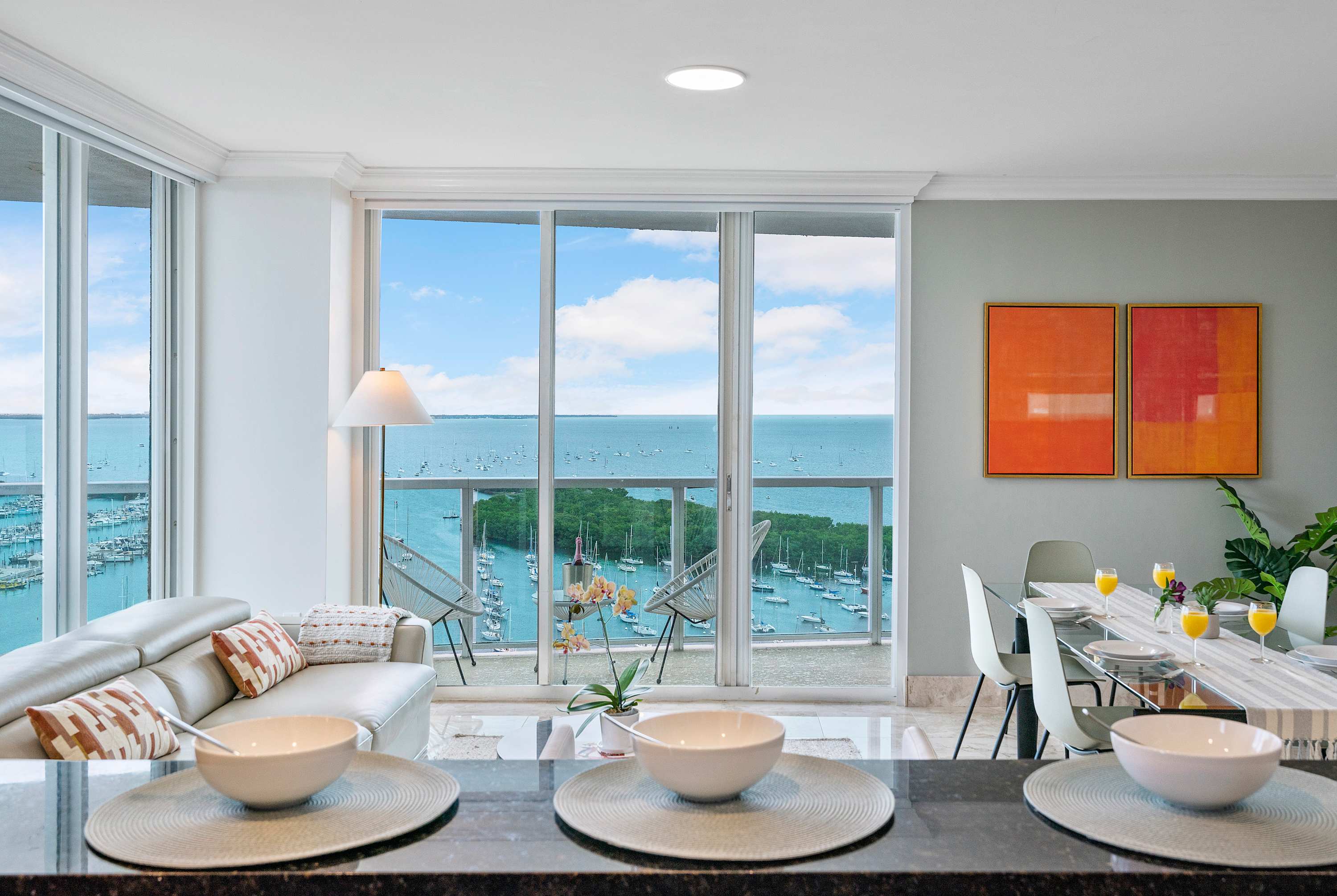 View from the kitchen into the open-concept living and dining area, featuring a glass dining table, a white sofa, and floor-to-ceiling windows offering stunning ocean and marina views.