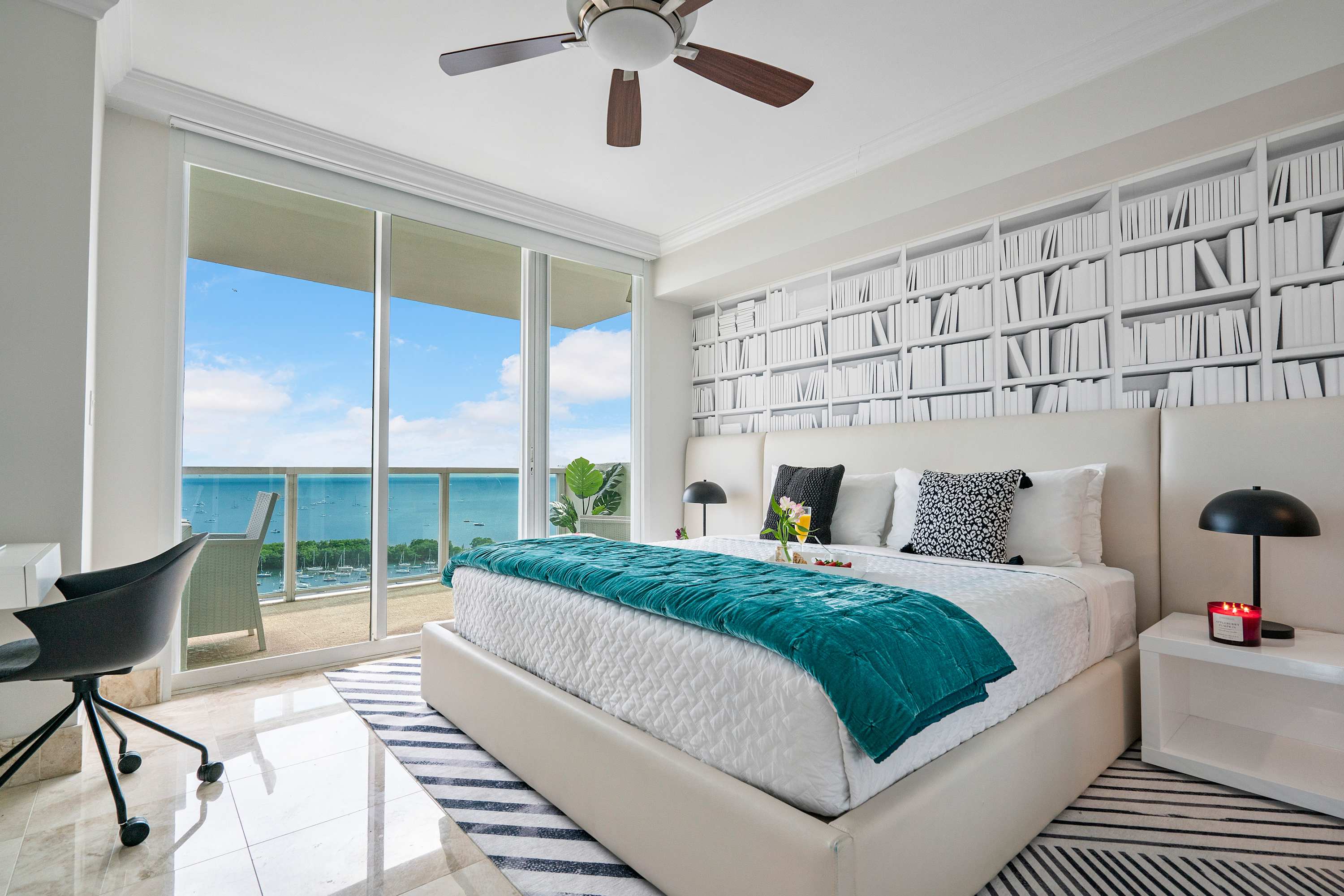 Spacious second bedroom with a large bed, a bookshelf-patterned accent wall, and a ceiling fan, featuring floor-to-ceiling glass doors leading to a balcony with bay views.