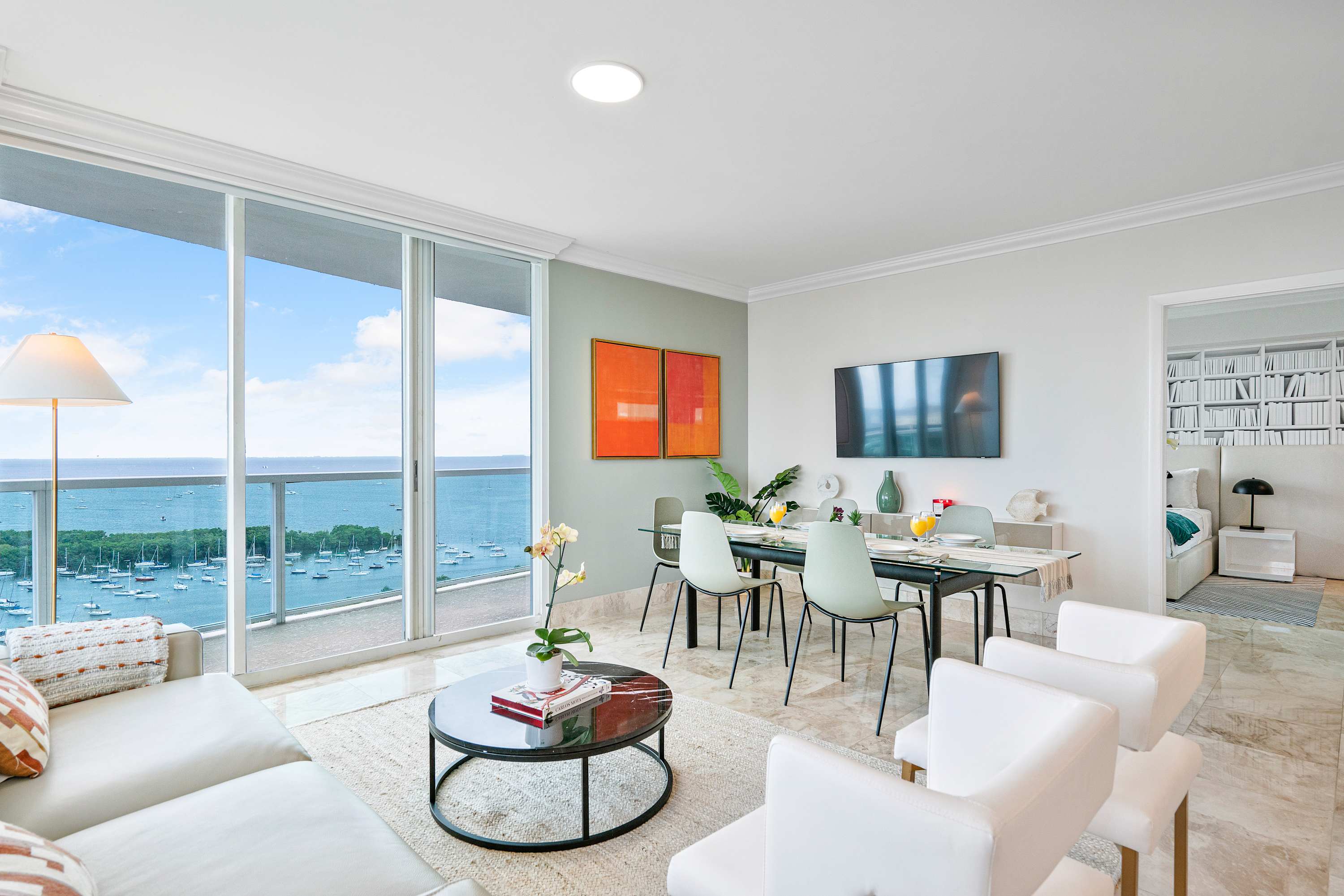 Open-concept living and dining area with light marble floors, a glass dining table, a white sofa, and floor-to-ceiling windows showcasing expansive ocean and marina views.
