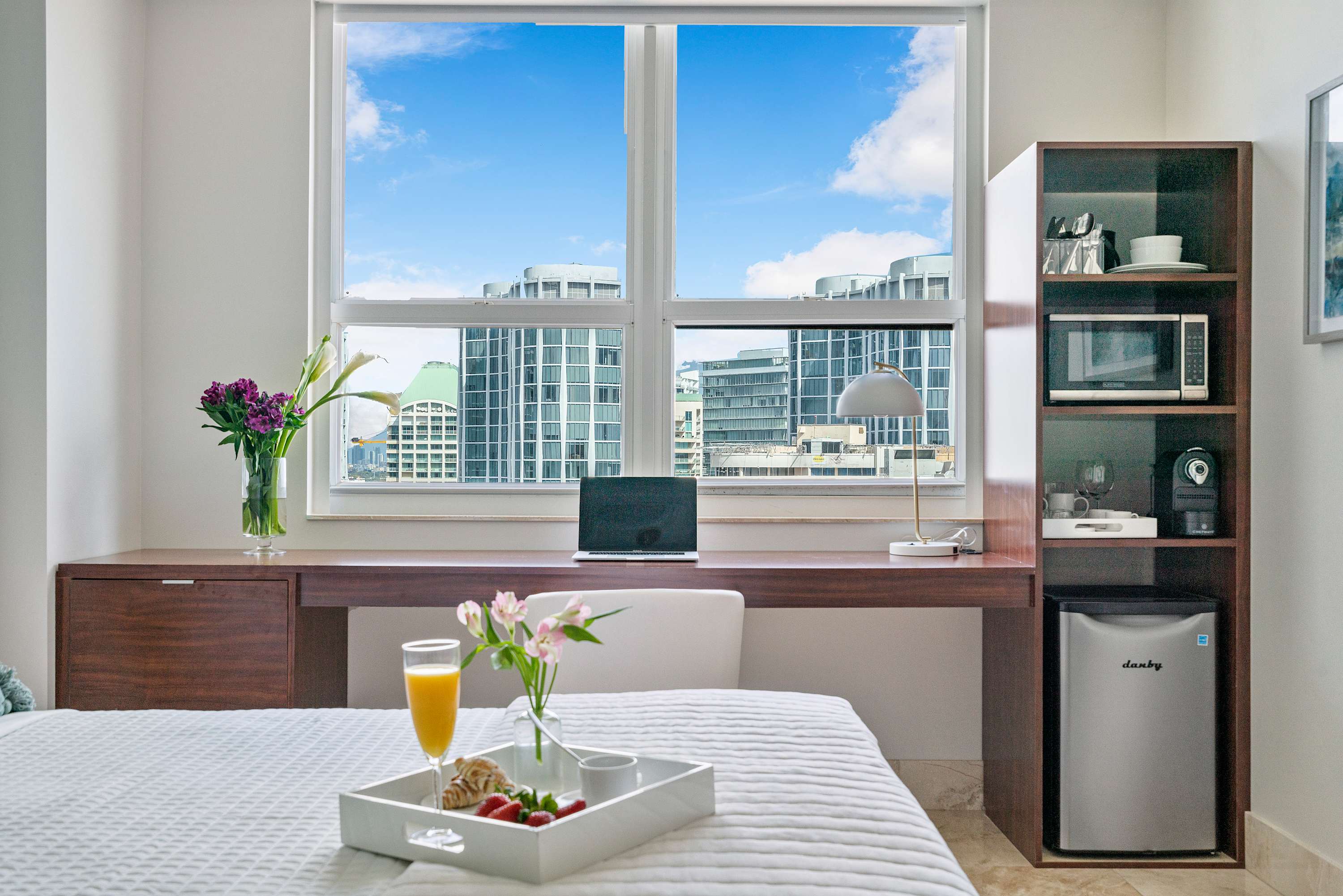 Close-up of the desk area in Bedroom 1, featuring a wooden desk with a laptop, a mini-fridge, and shelving unit, with city buildings visible through the large window.