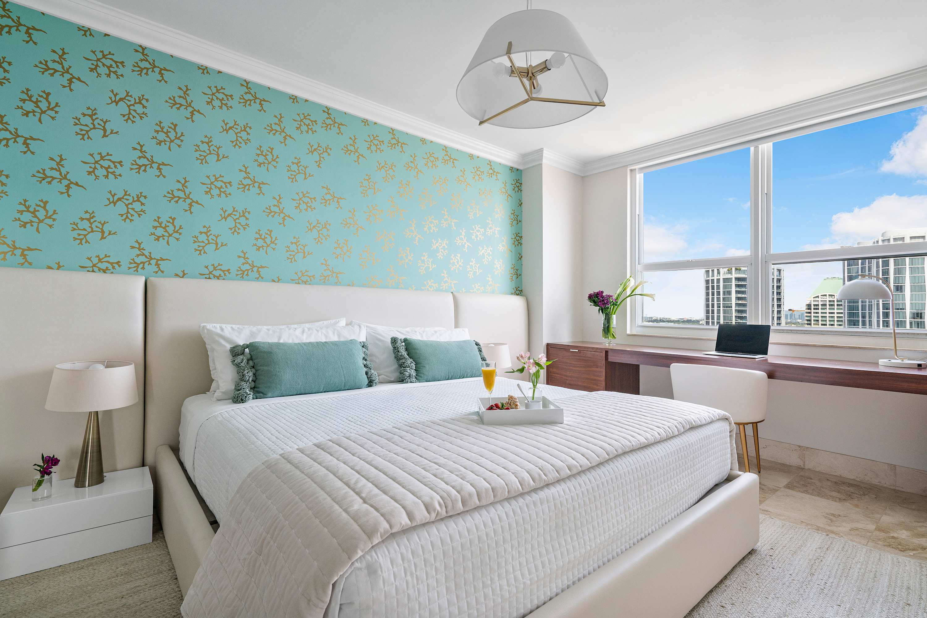 Bright bedroom with a white bed, a teal accent wall with gold coral patterns, and a built-in desk area with a laptop, offering city views through a large window.