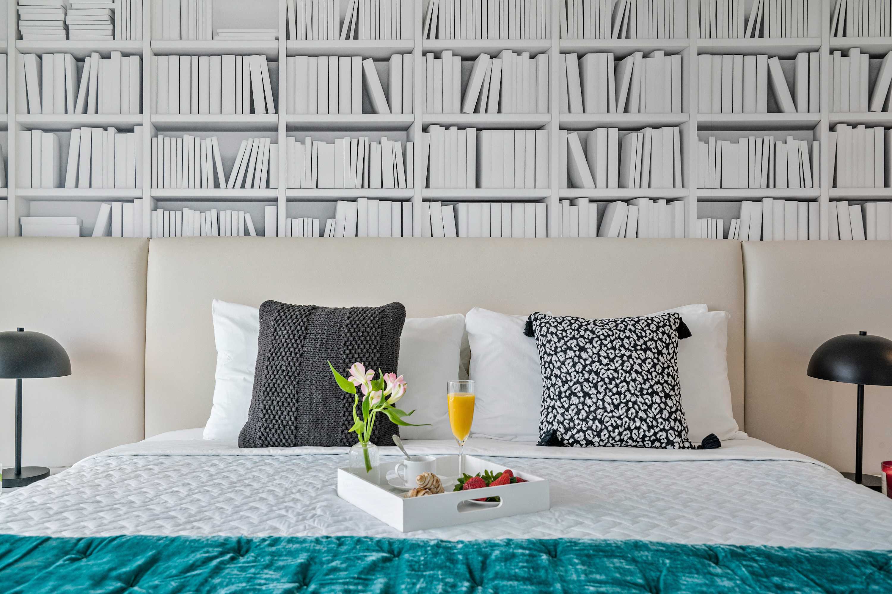 Second bedroom featuring a large bed with a teal throw blanket, a unique bookshelf-patterned accent wall, and black and white decorative pillows, with a breakfast tray on the bed.