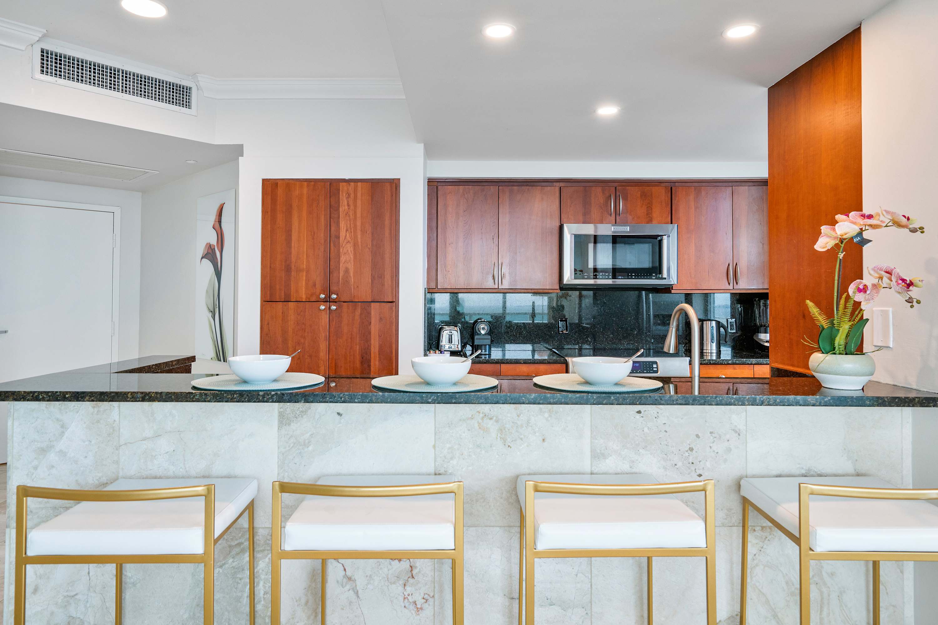 Modern kitchen with dark wood cabinetry, black granite countertops, and a breakfast bar with four white and gold bar stools. Stainless steel appliances and recessed lighting complete the sleek design.