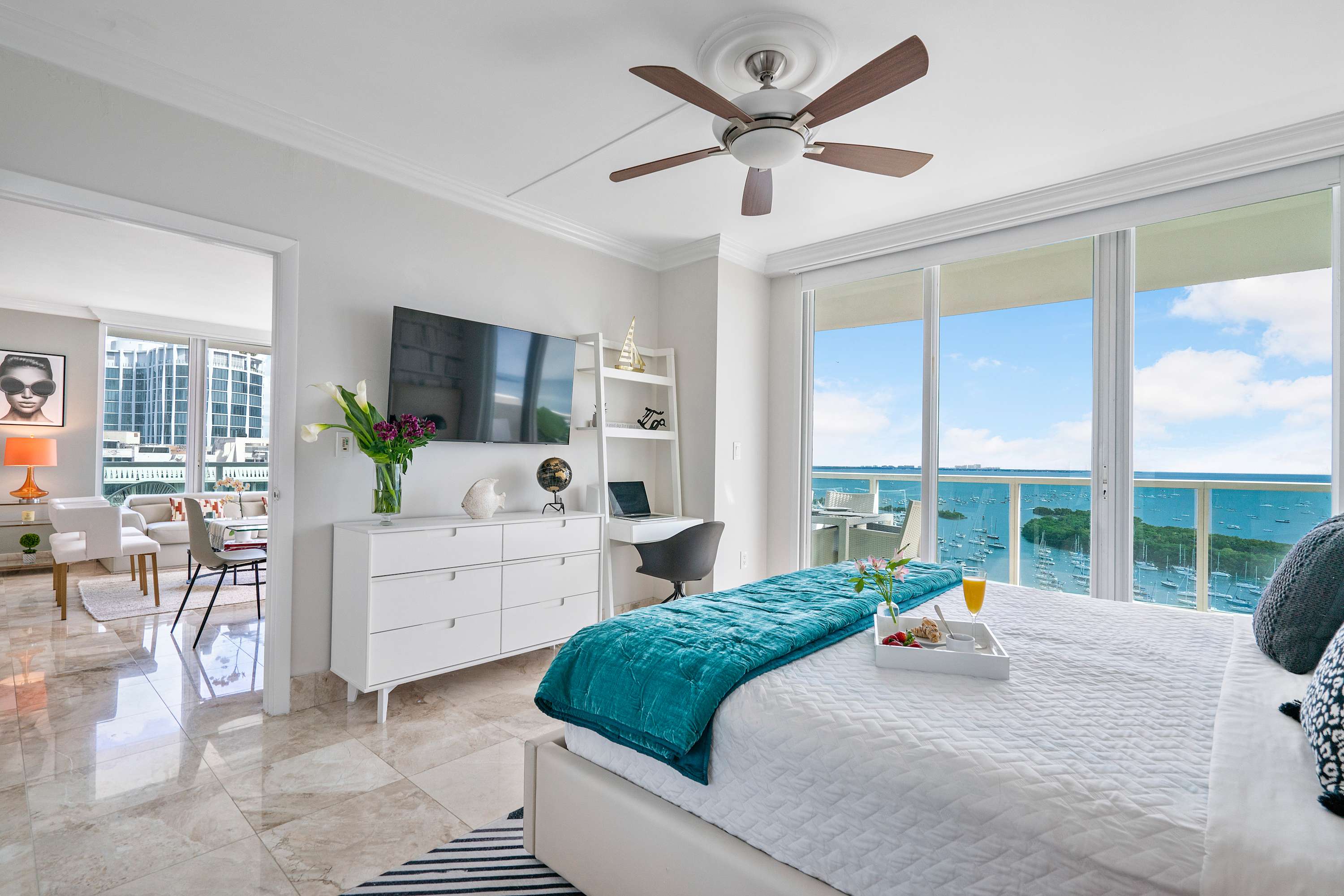 Bright bedroom with a white bed, teal throw blanket, and a tray with breakfast, featuring a ceiling fan, a wall-mounted TV, a desk, and floor-to-ceiling windows with stunning ocean and marina views.