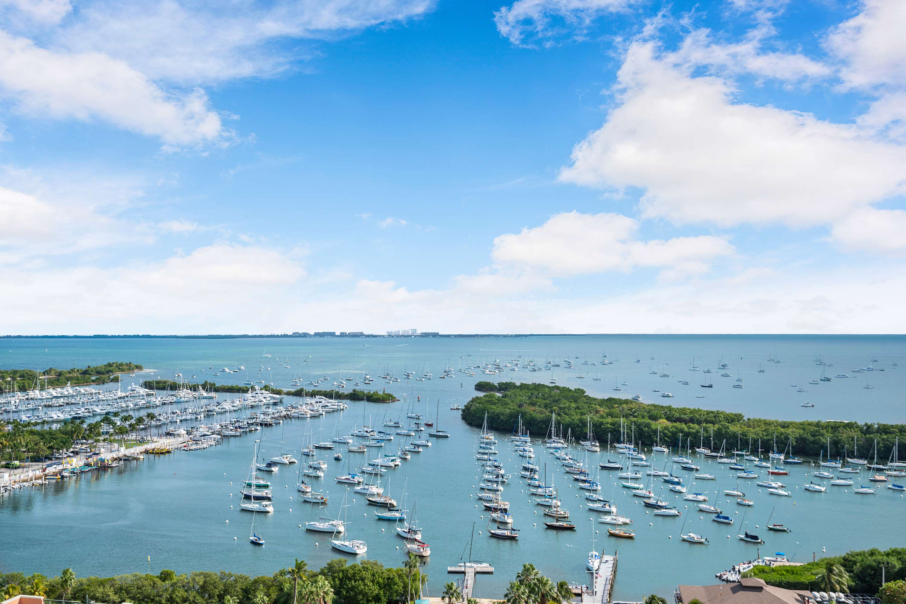 Expansive aerial view of a vibrant marina filled with numerous sailboats and yachts, surrounded by lush green islands and the vast blue ocean under a partly cloudy sky.