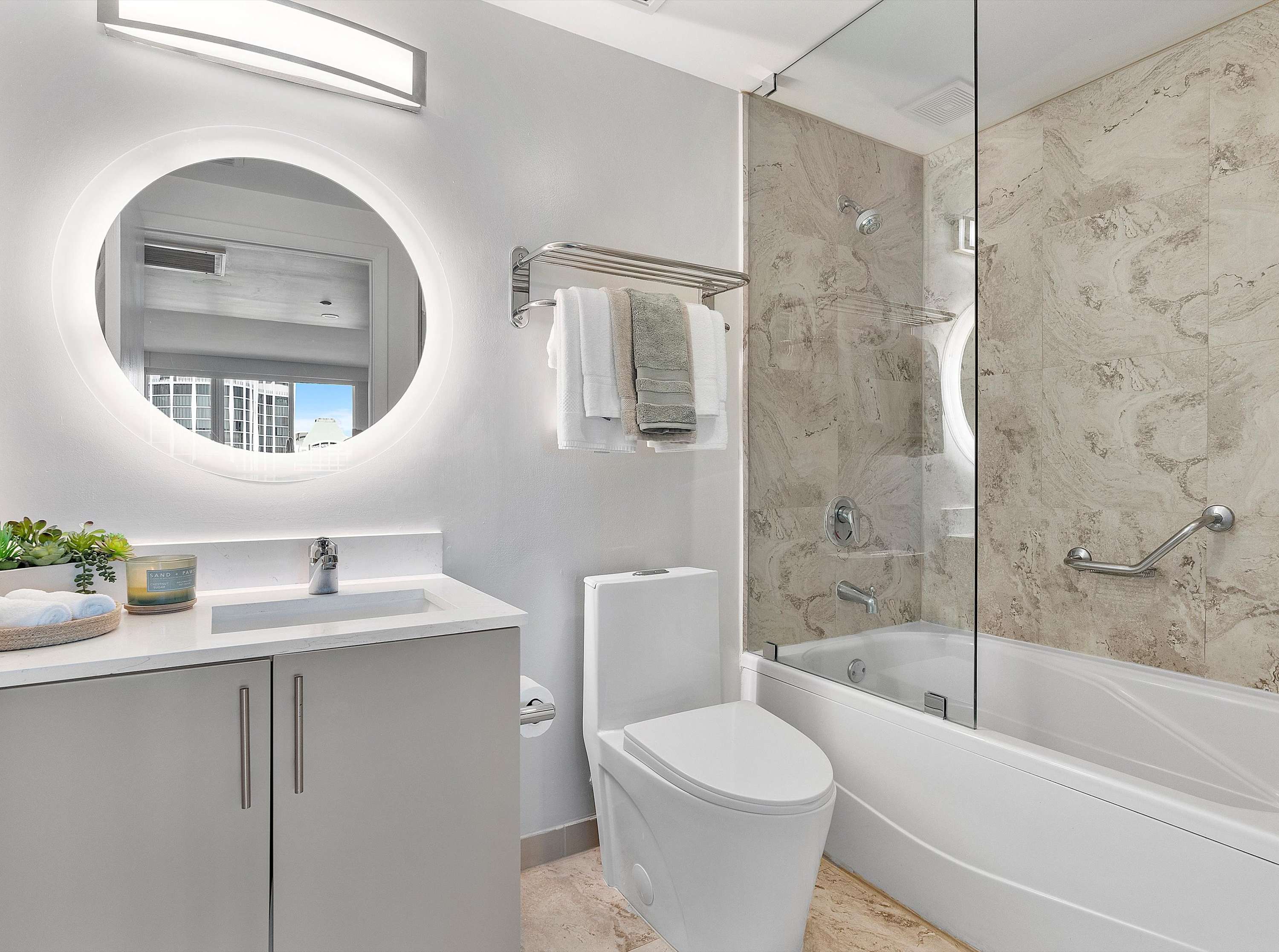 Bright, contemporary bathroom with a white vanity, a round illuminated mirror, and a toilet beside a bathtub with a glass partition. The shower area has light marble-patterned tiles, and a towel rack holds neatly folded towels.
