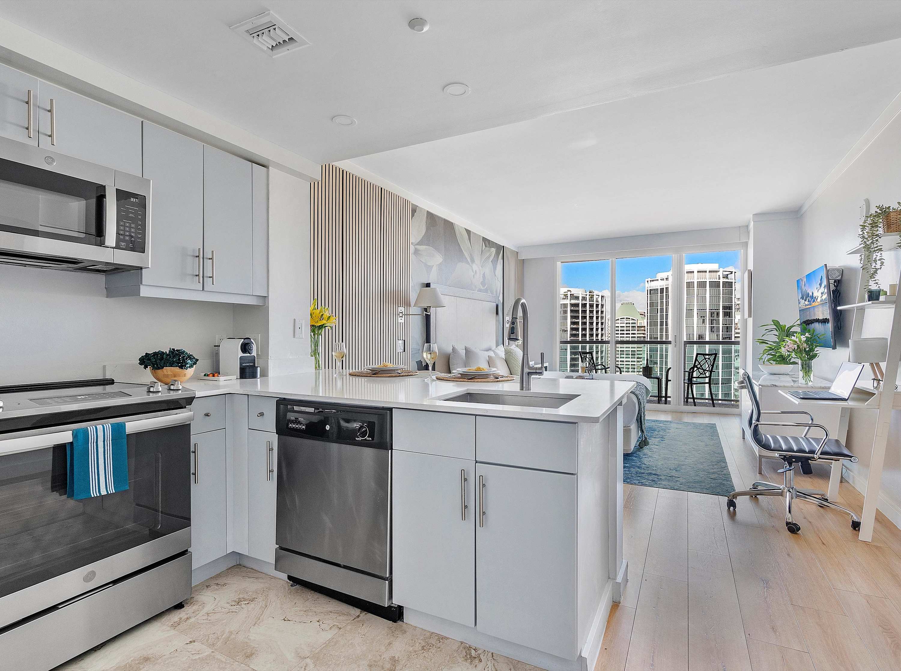 A bright, open-concept kitchen featuring light gray cabinetry, stainless steel appliances including a refrigerator, oven, and dishwasher, and a white quartz island with bar seating, seamlessly connecting to the living space.